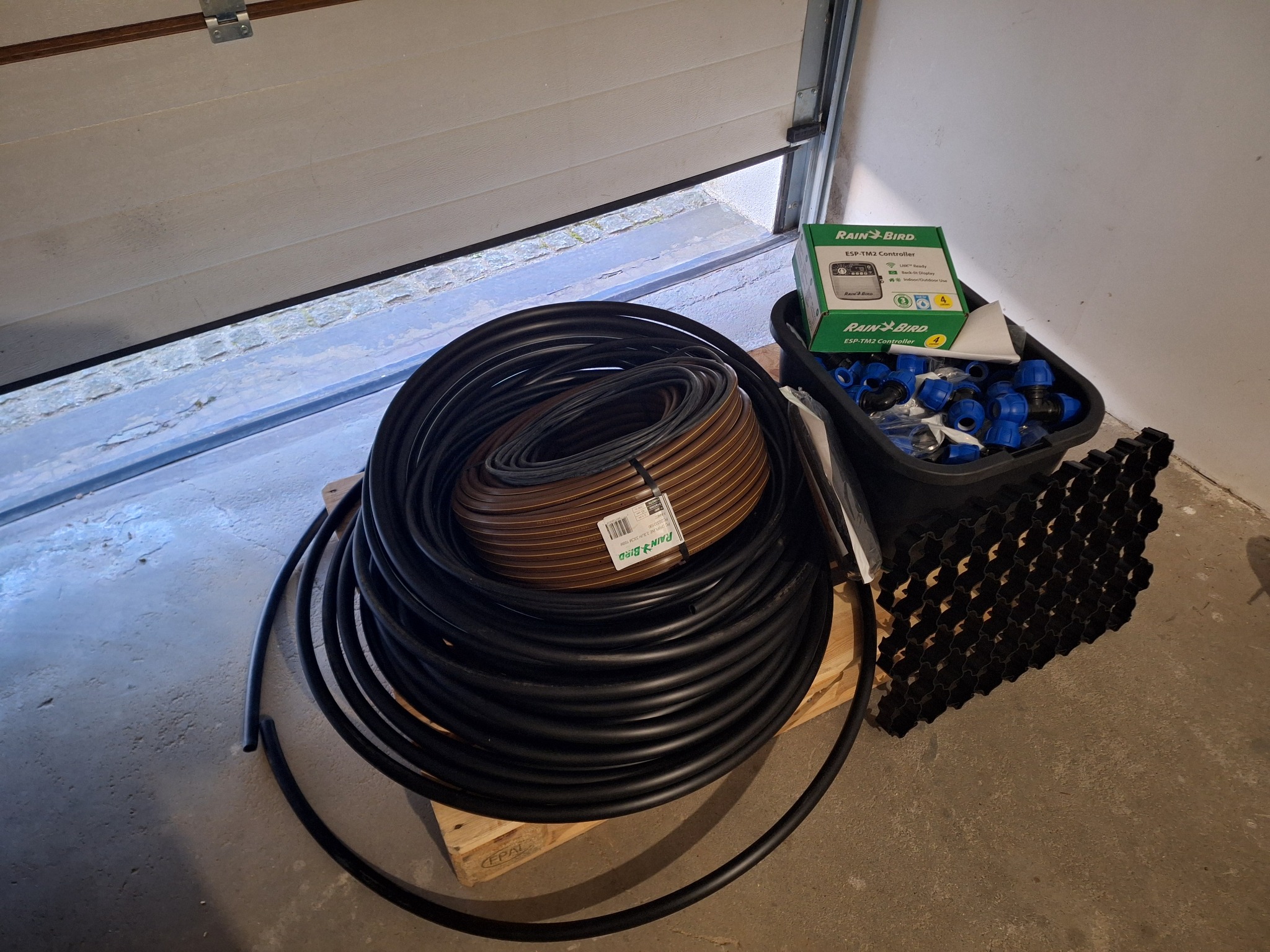Zestaw elementów do instalacji nawadniania: zwój czarnego węża nawadniającego, brązowy wąż kroplujący, sterownik Rain Bird ESP-TM2, złączki i kratka trawnikowa, ułożone na palecie w garażu.