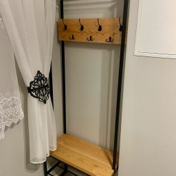 OSTRY SPAW - Metalowy wieszak na ubrania z drewnianym siedziskiem i półką, widoczne haczyki i dekoracyjna zasłona z ornamentem.