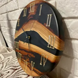 Okrągły zegar z drewna i czarnej żywicy epoksydowej, z rzymskimi cyframi w kolorze złotym, umieszczony na koronkowej serwecie i tle z białej cegły.