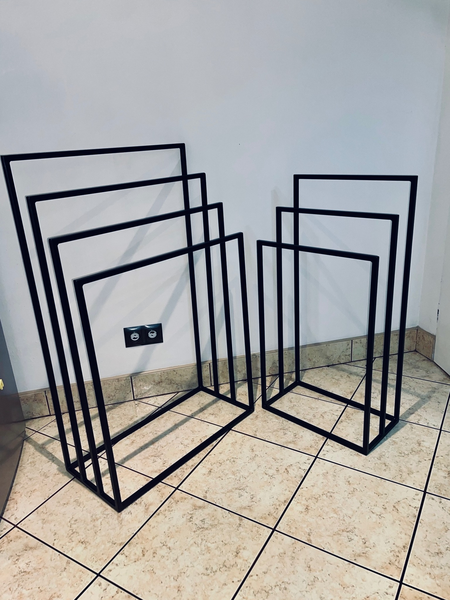 Dwie grupy czarnych, metalowych, kwadratowych ram o różnych rozmiarach, ustawione na kafelkach podłogowych w pobliżu białej ściany z gniazdkiem elektrycznym.