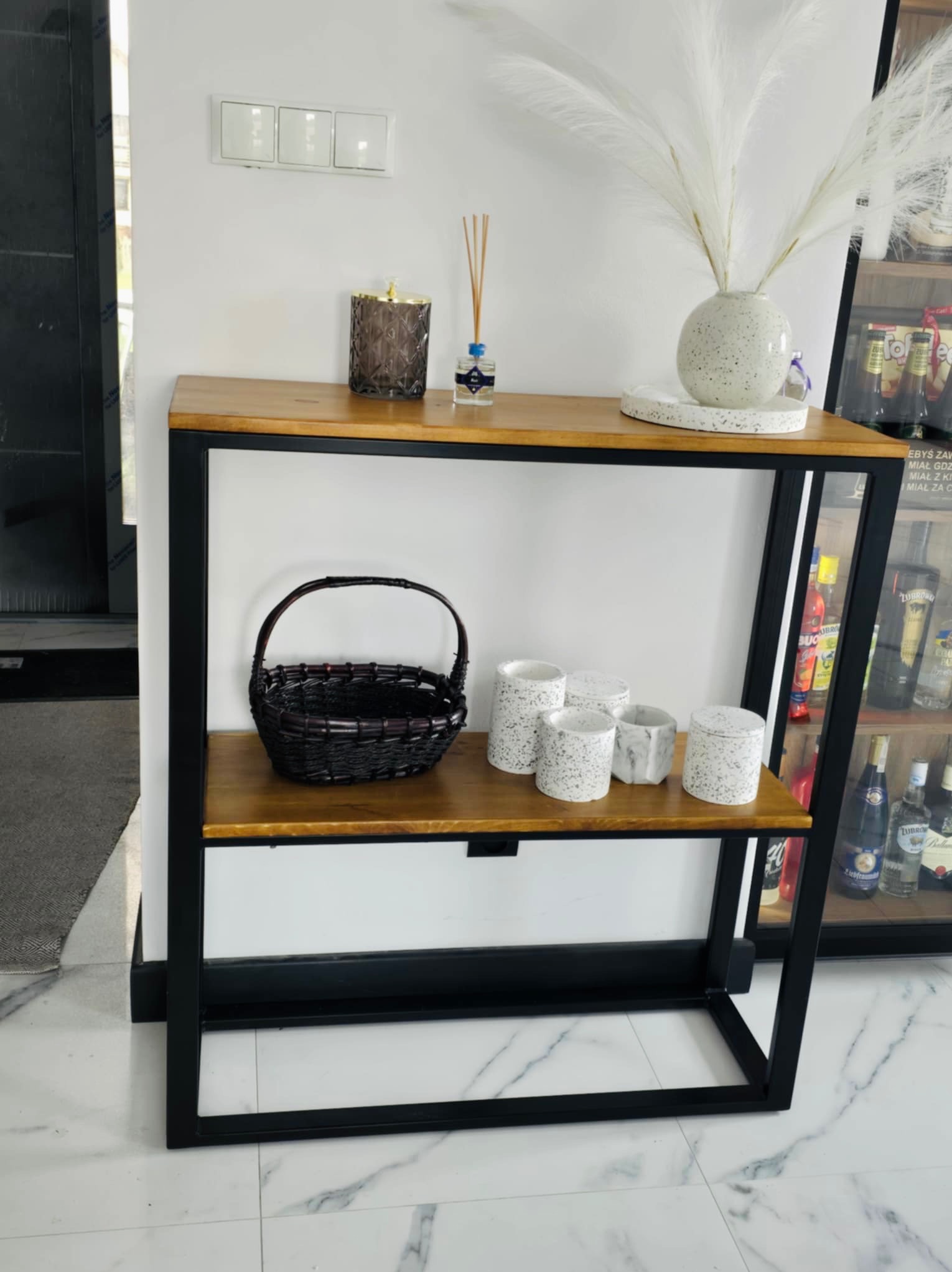 Konsola z drewnianym blatem i metalową ramą, na której ustawione są dekoracje: koszyk, ceramiczne doniczki i wazon z suchymi trawami, w tle przeszklona witryna z alkoholem.