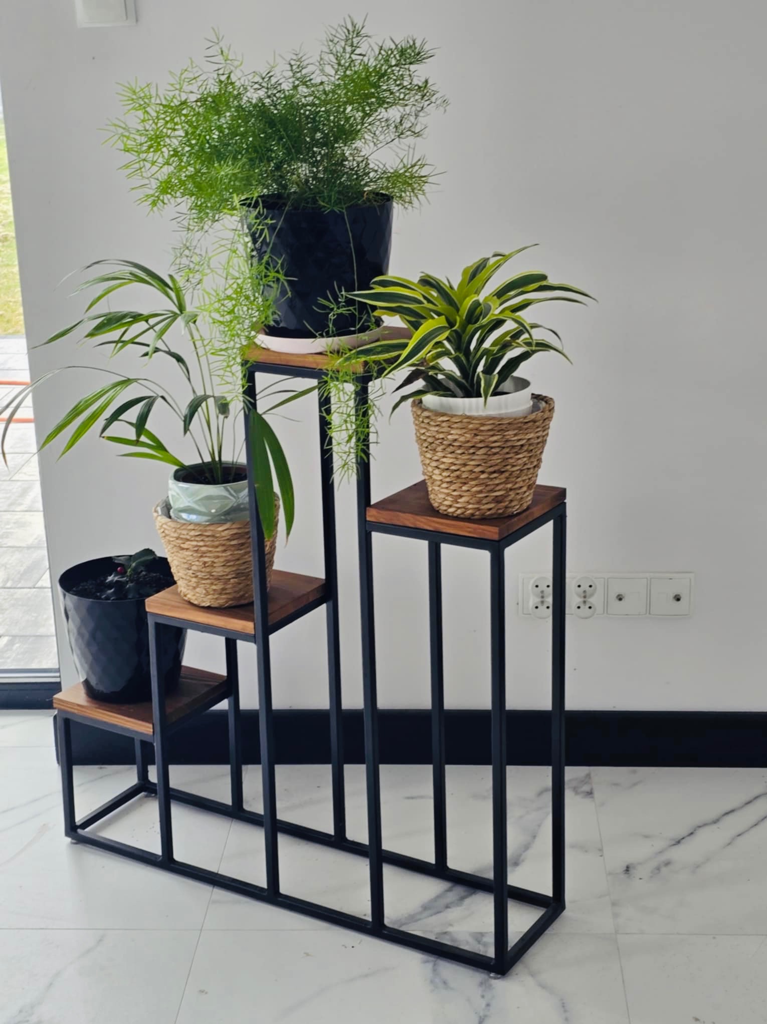 Metalowy kwietnik z drewnianymi półkami, na których stoją cztery różne rośliny doniczkowe, na tle białej ściany i marmurowej podłogi.