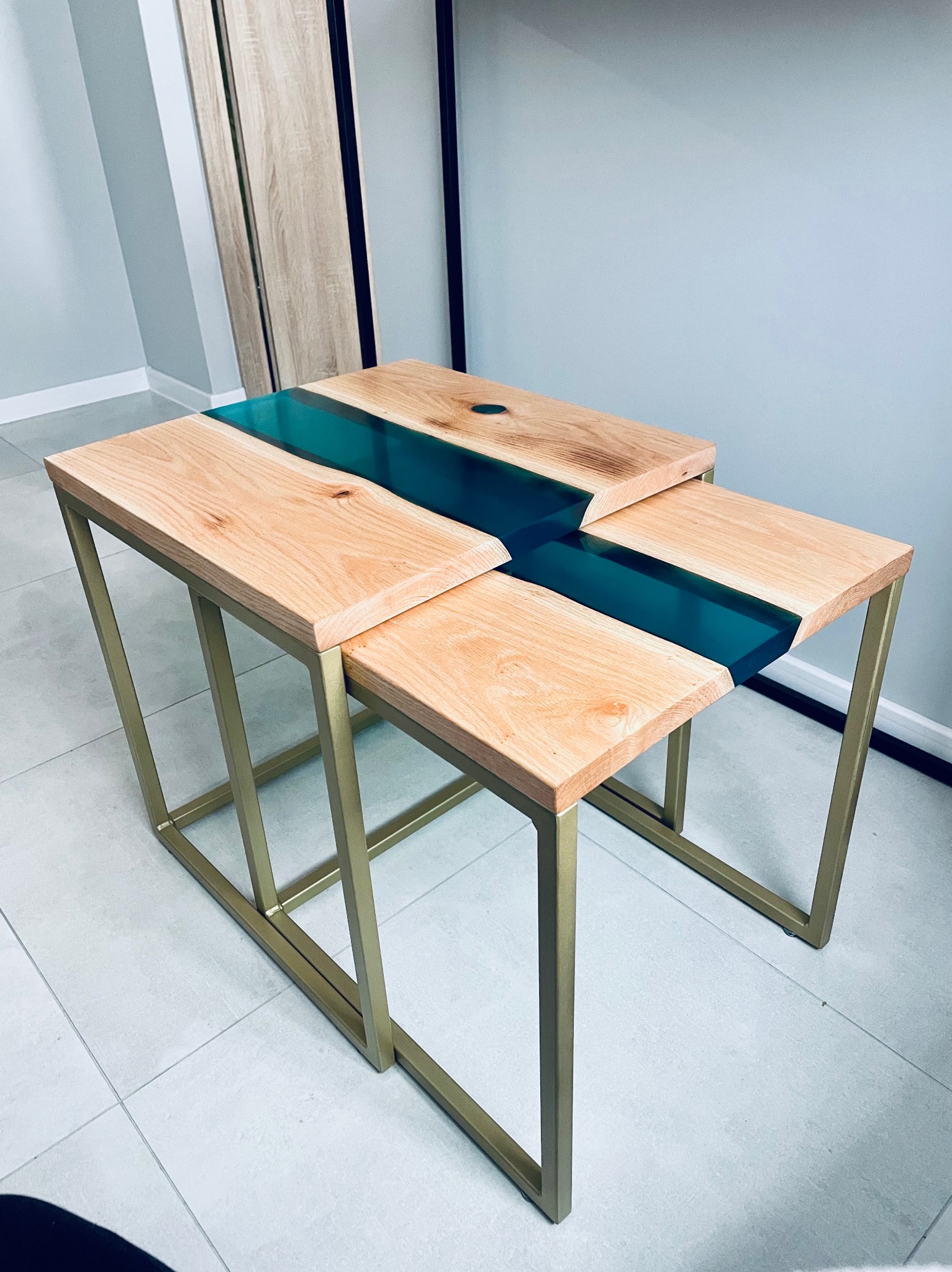 Zestaw dwóch stolików kawowych z drewnianymi blatami i złotymi metalowymi nogami, wypełnionych turkusową żywicą epoksydową, ustawionych jeden pod drugim.