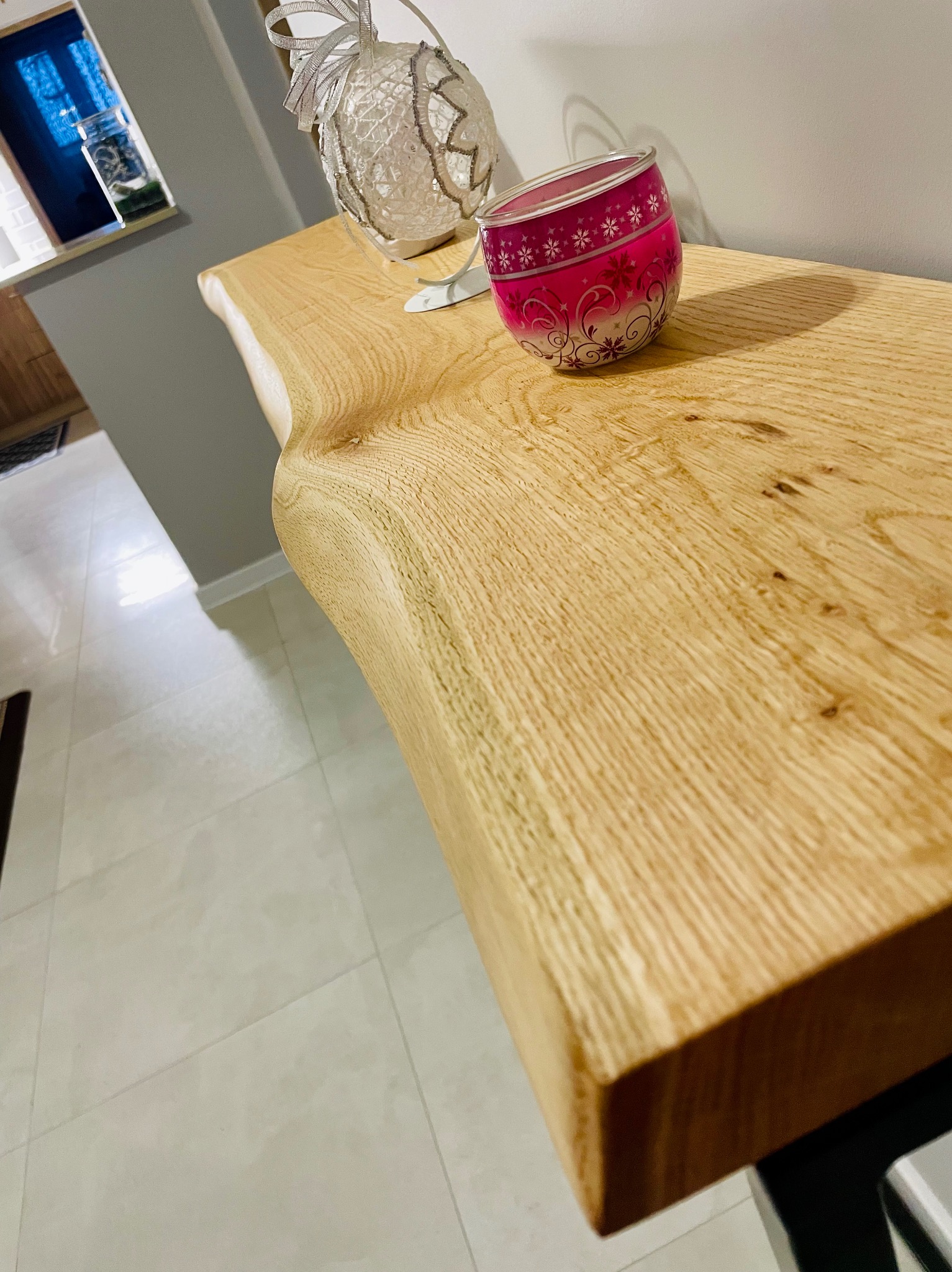 Blat drewniany o nieregularnym kształcie krawędzi, z dekoracjami w postaci białej kuli z kokardą i różowego świecznika, ustawiony w nowoczesnym wnętrzu.