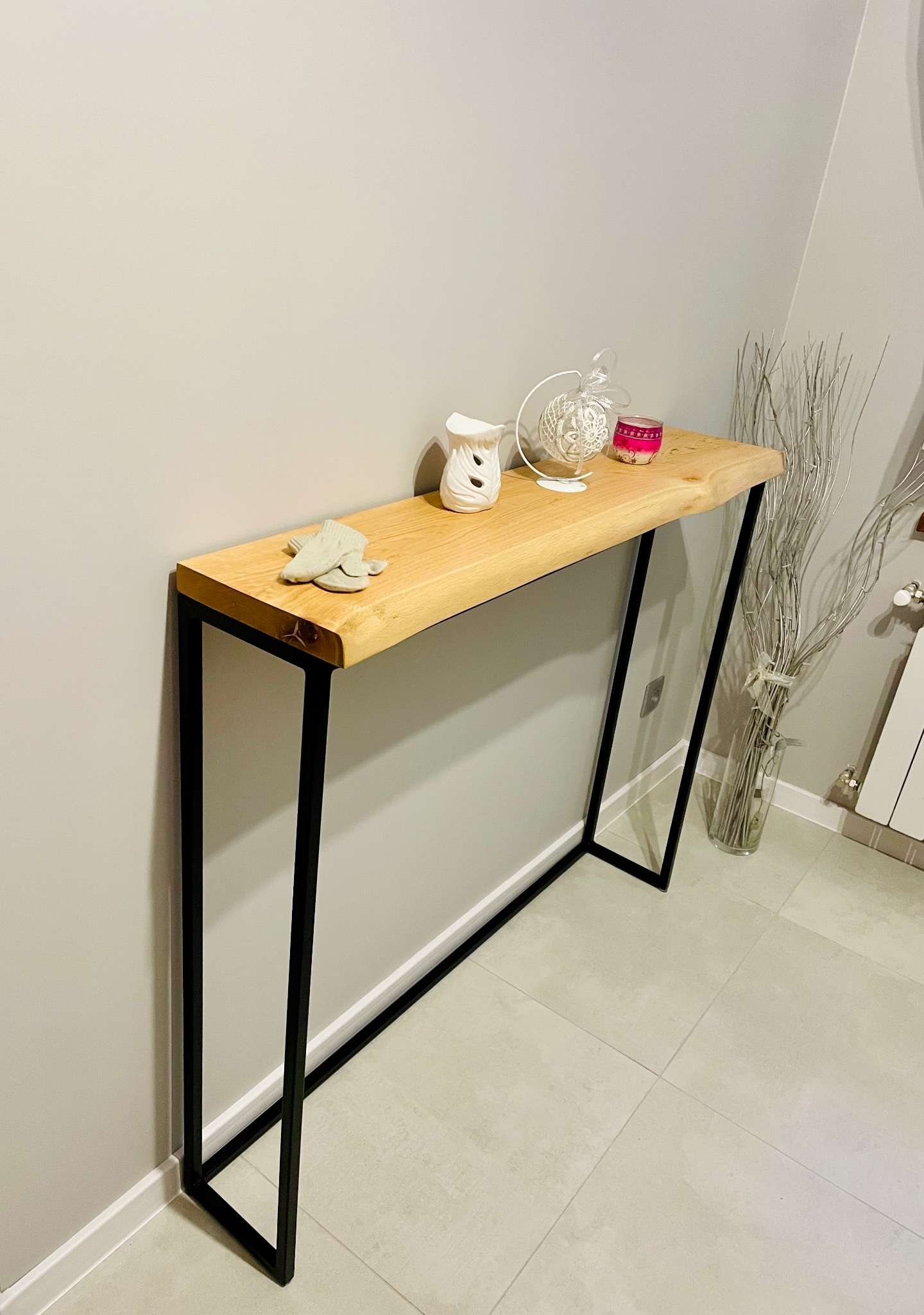 Wąski stolik konsola z blatem z jasnego drewna i czarnymi metalowymi nogami, dekorowany ceramiczną ozdobą, szklanym wazonem z dekoracyjnymi gałązkami i parą rękawiczek.