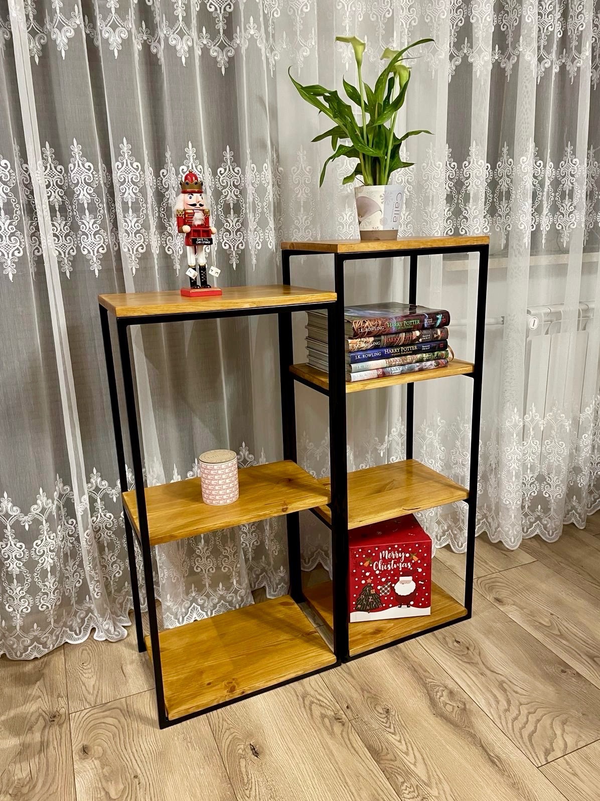 Regał z drewnianymi półkami i czarną metalową ramą, na którym znajdują się książki, kwiaty w doniczce, ozdobny dziadek do orzechów i pudełko z motywem świątecznym, na tle białych, wzorzystych zasłon.
