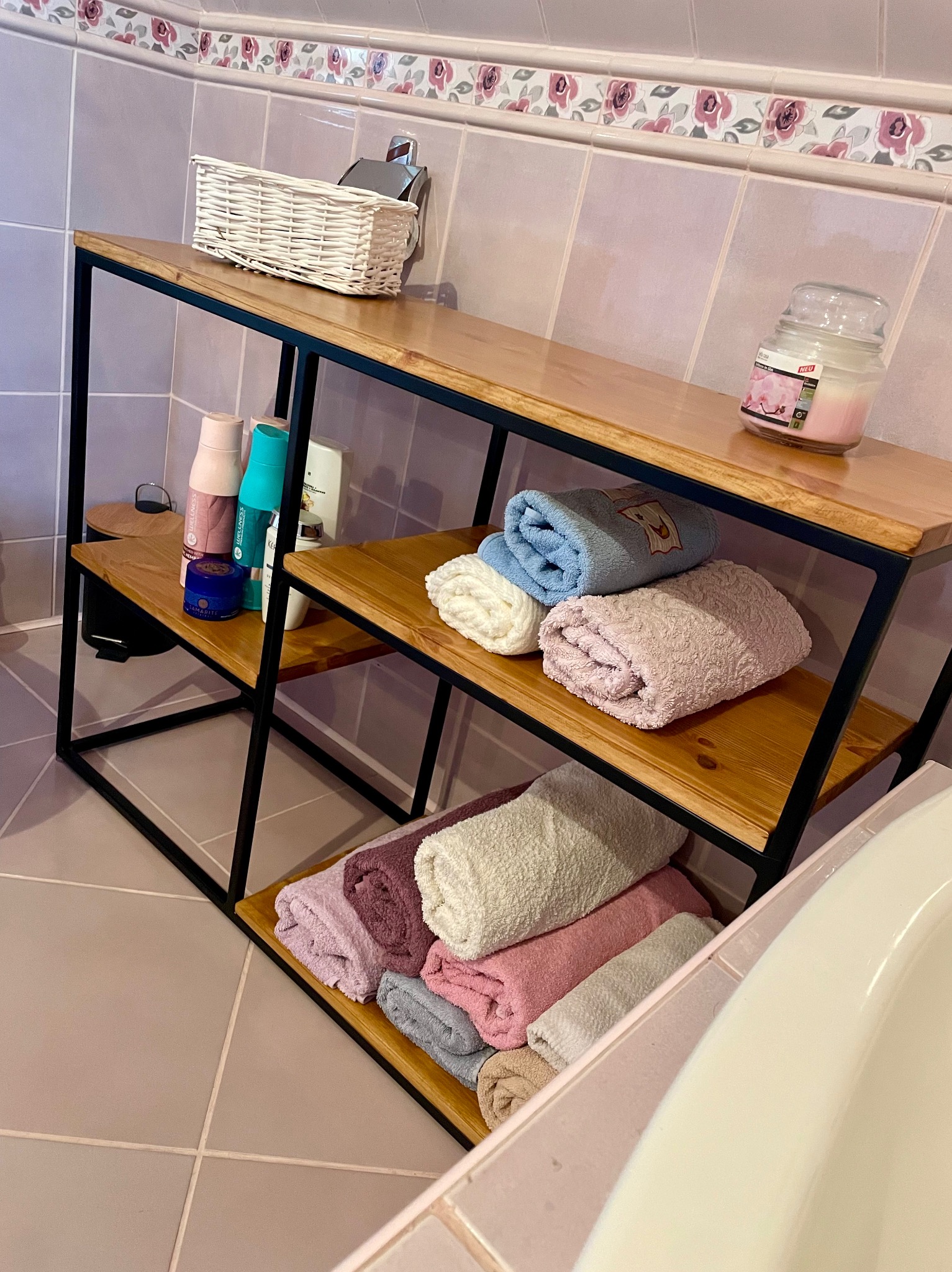 Metalowy regał z drewnianymi półkami w łazience, na którym ułożone są ręczniki w różnych kolorach oraz kosmetyki i świeca zapachowa.