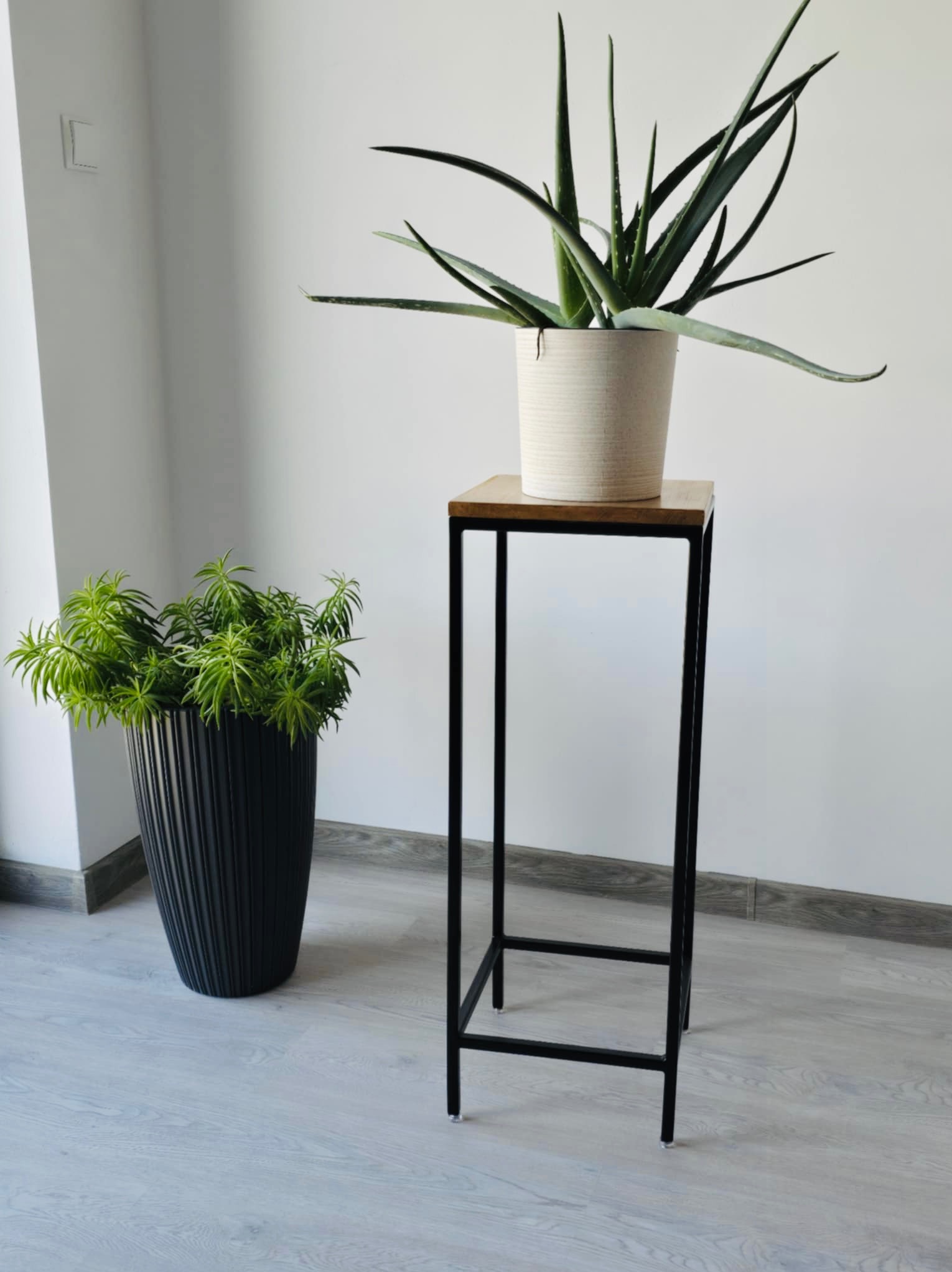 Metalowy kwietnik w stylu loft z drewnianym blatem, na którym stoi aloes w beżowej doniczce, obok czarna donica z zieloną rośliną, na tle jasnej ściany i podłogi.