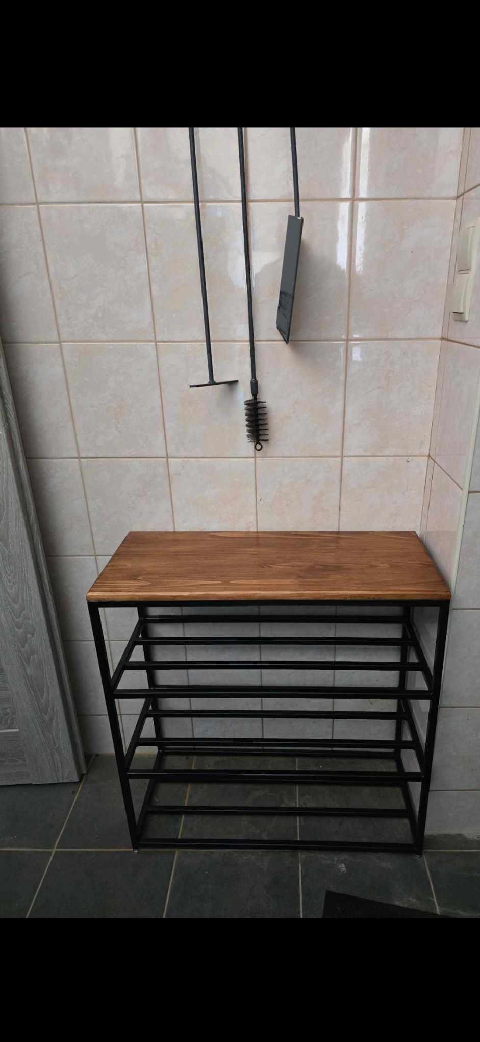 Metalowy regał na buty z drewnianym blatem, stojący na podłodze z płytek, z akcesoriami kominkowymi wiszącymi na ścianie w tle.