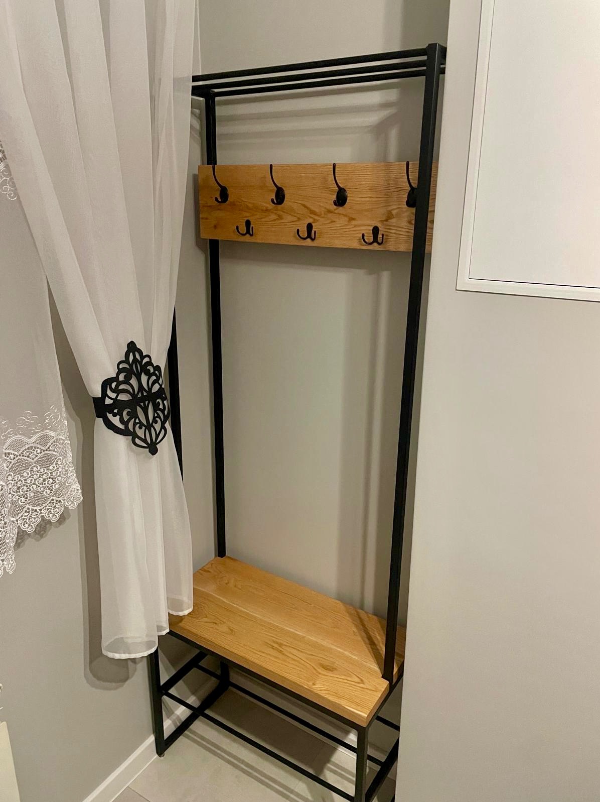 Metalowy wieszak na ubrania z drewnianym siedziskiem i półką, widoczne haczyki i dekoracyjna zasłona z ornamentem.