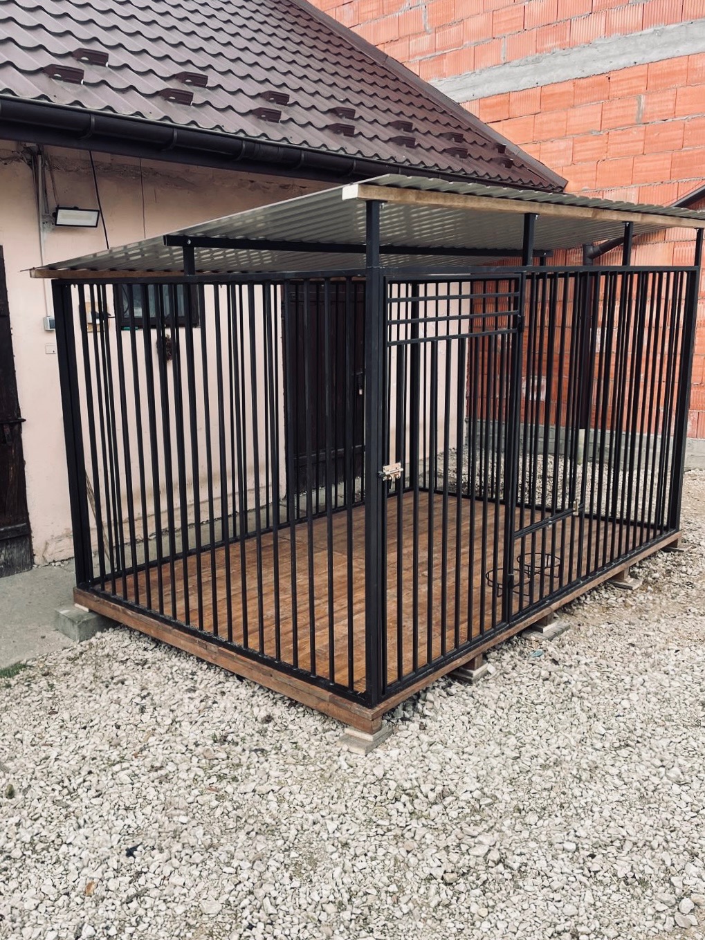 Metalowa konstrukcja z zadaszeniem i drewnianą podłogą, prawdopodobnie kojec dla psa, ustawiona na zewnątrz na żwirowym podłożu, z budynkiem w tle.