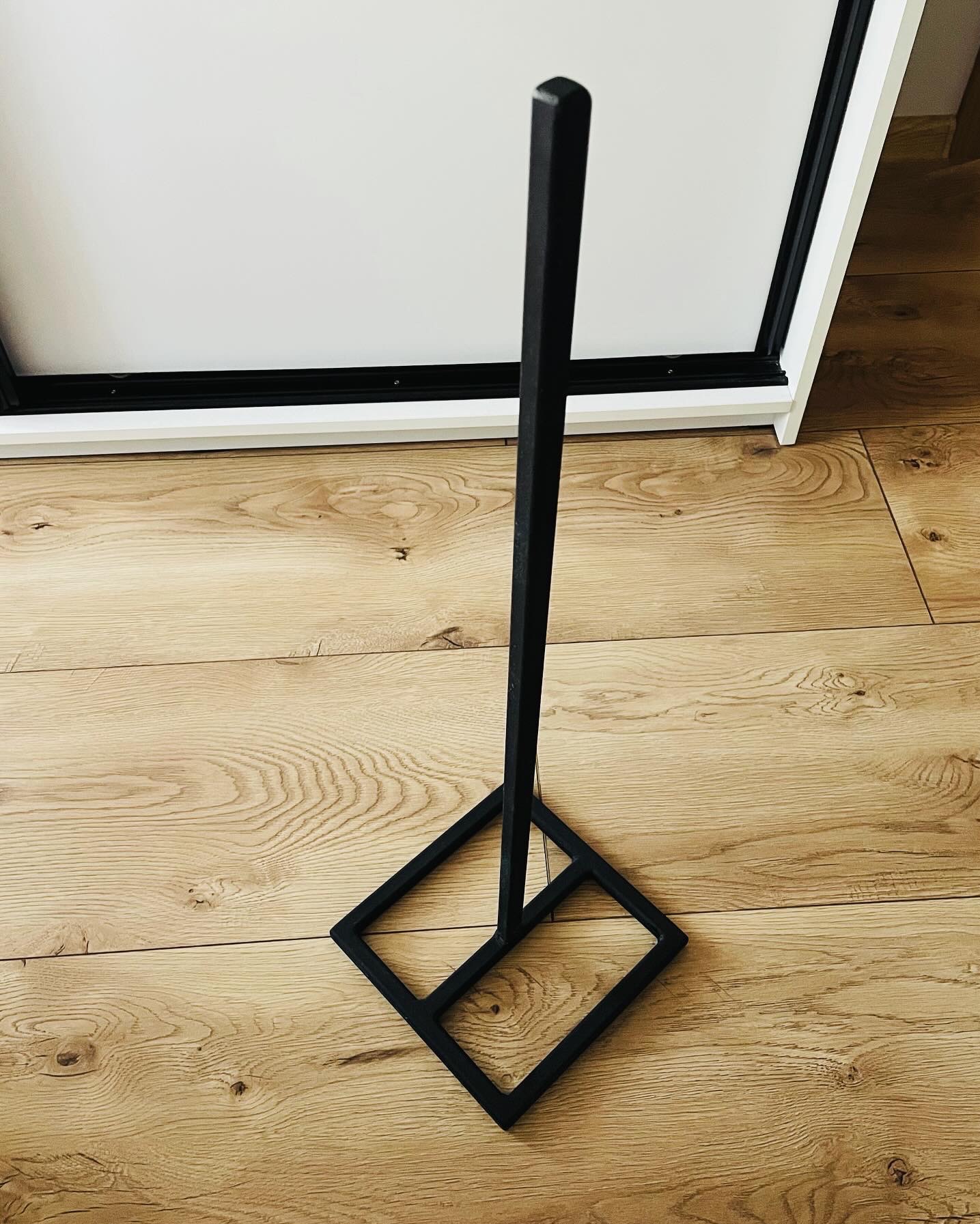 Metalowy stojak o kwadratowej podstawie i pionowym słupku, ustawiony na drewnianej podłodze, z fragmentem białej szafy w tle.