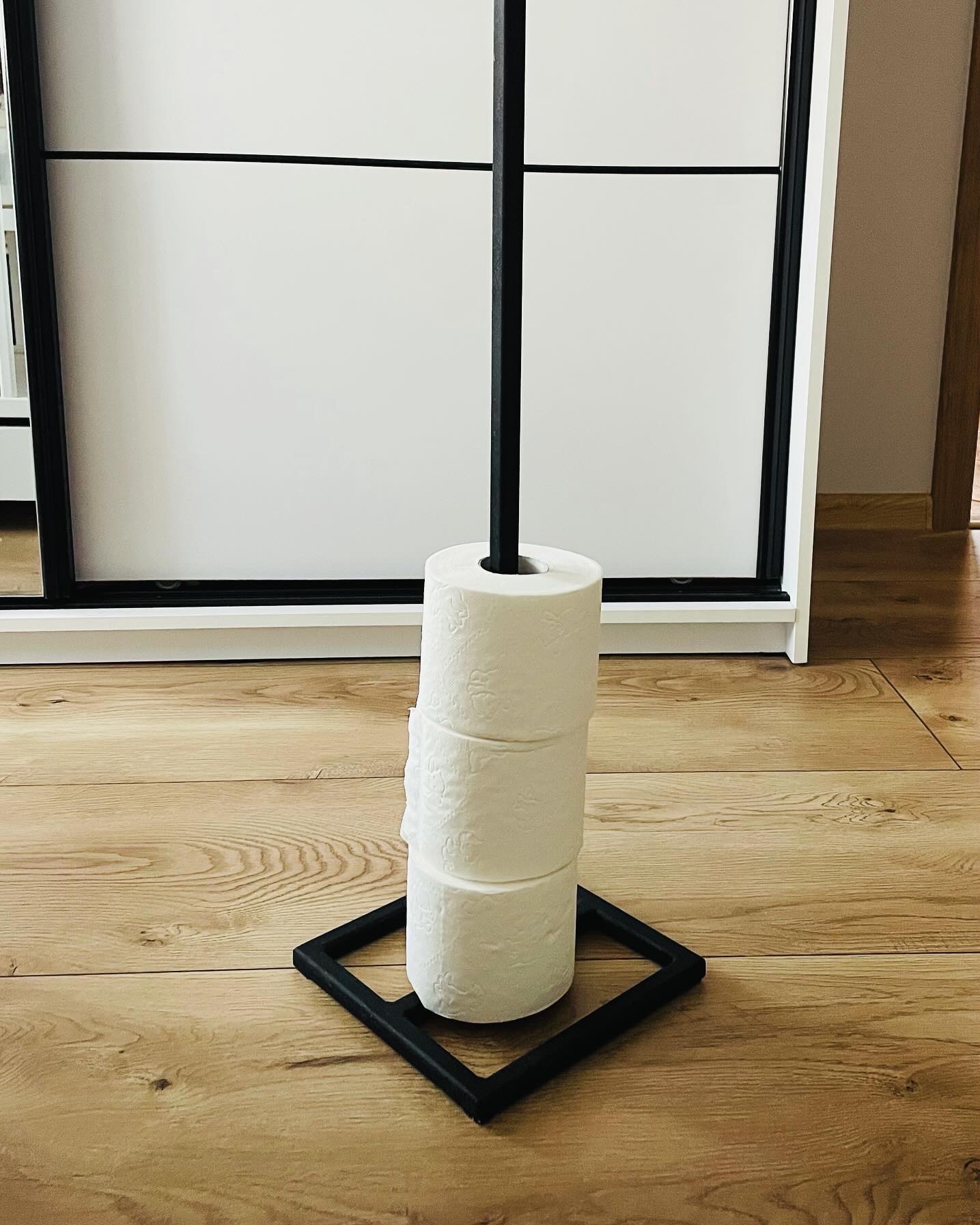 Stojak na papier toaletowy z czarną, metalową podstawą w kształcie kwadratu i pionowym prętem, na którym umieszczone są trzy rolki białego papieru toaletowego; w tle widoczna biała szafa przesuwna...