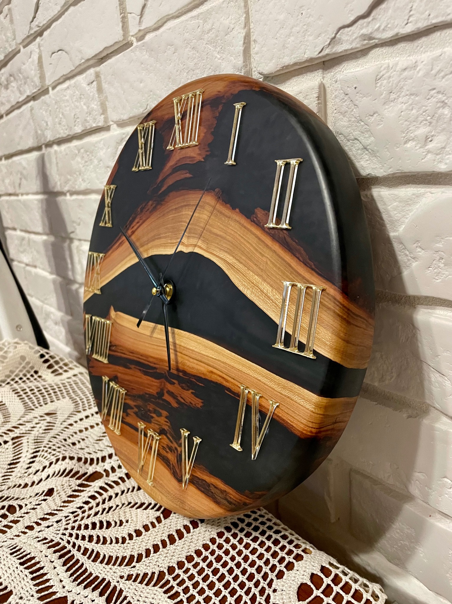 Okrągły zegar z drewna i czarnej żywicy epoksydowej, z rzymskimi cyframi w kolorze złotym, umieszczony na koronkowej serwecie i tle z białej cegły.