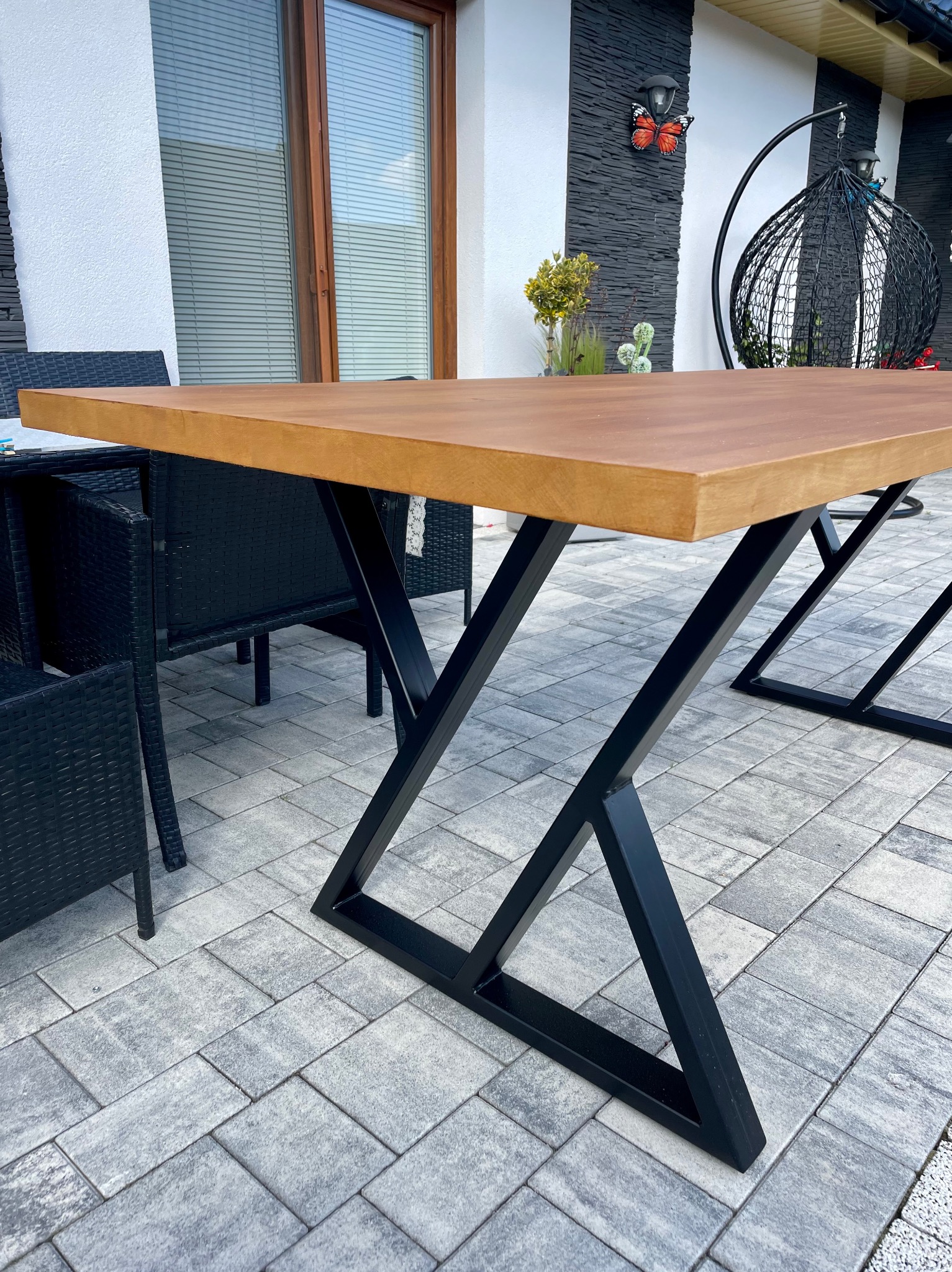 Stół z drewnianym blatem i czarnymi, metalowymi nogami w kształcie litery X. W tle czarne krzesła z rattanu i dekoracje na ścianie.