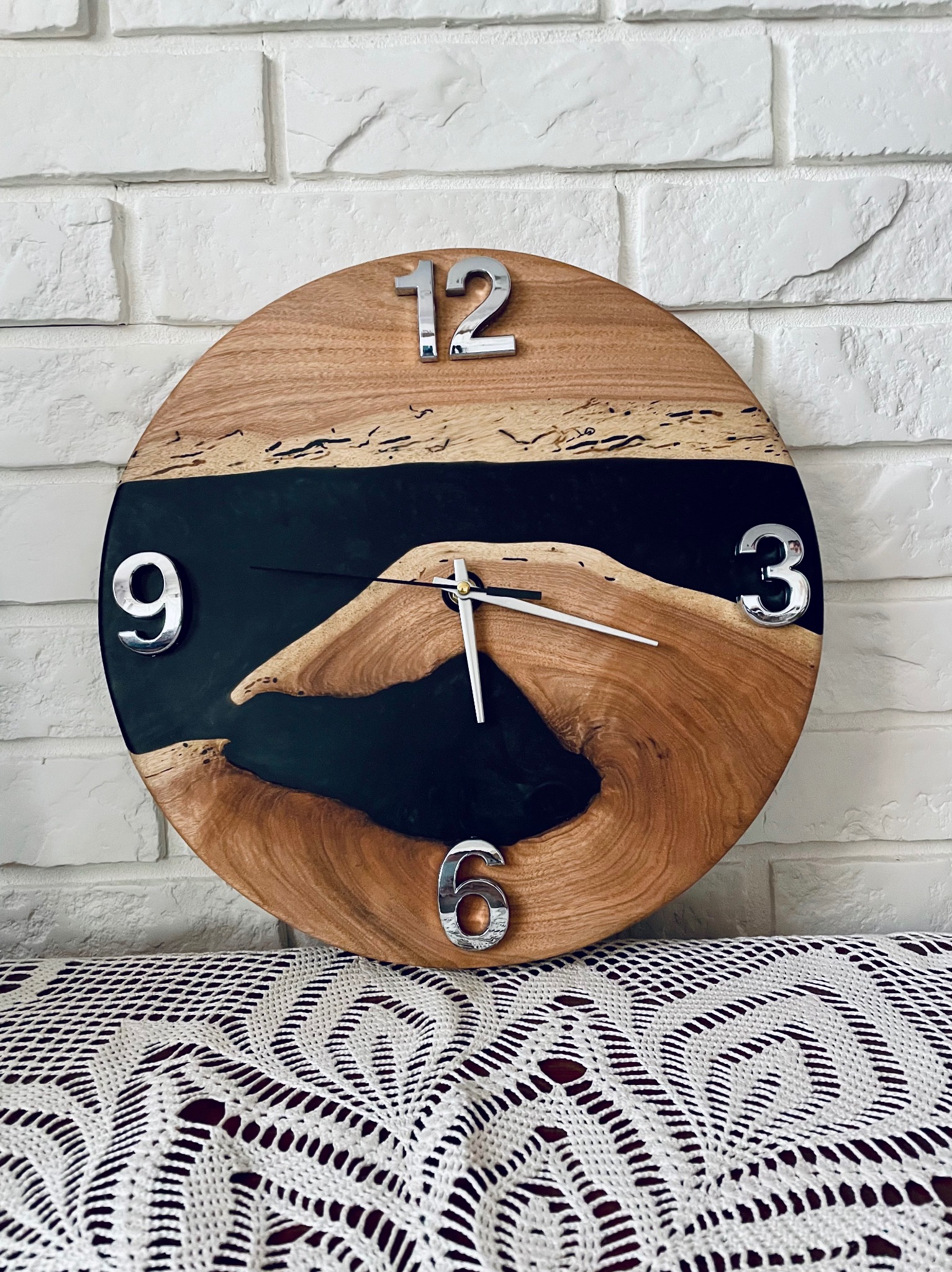 Okrągły zegar z drewna i żywicy epoksydowej, cyfry 3D w kolorze chromu, na tle białej cegły i koronkowej serwety. Unikalny design.