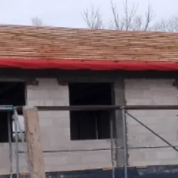 Wznoszenie ścian budynku z bloczków, widoczne okna bez stolarki, drewniana konstrukcja dachu z czerwoną membraną, rusztowanie wzdłuż ściany.