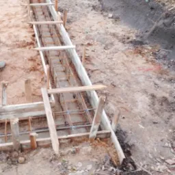 Przygotowanie szalunku i zbrojenia pod fundament liniowy, widoczne drewniane deski i stalowe pręty zbrojeniowe, ziemia i trawa w tle.