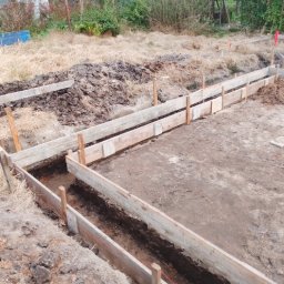 Rafał Rot - Wykonanie szalunku pod fundamenty z drewnianych desek, widoczne wykopane rowy, pionowe pale podtrzymujące konstrukcję i ziemia usypana obok wykopu.