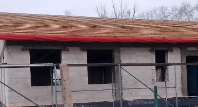 Wznoszenie ścian budynku z bloczków, widoczne okna bez stolarki, drewniana konstrukcja dachu z czerwoną membraną, rusztowanie wzdłuż ściany.