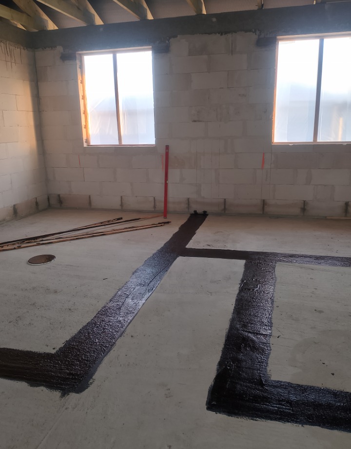 Wnętrze surowego domu w trakcie budowy z fundamentami zabezpieczonymi czarną hydroizolacją, ściany z białych bloczków, dwa okna z widocznym światłem dziennym, poziomica oparta o ścianę.