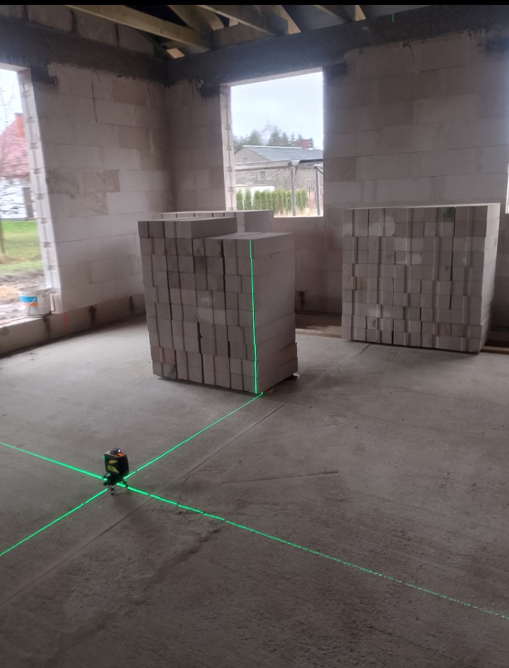 Wnętrze surowego domu w trakcie budowy z ustawionym laserem krzyżowym, palety z bloczkami betonu komórkowego, widoczne okna z widokiem na zewnątrz.