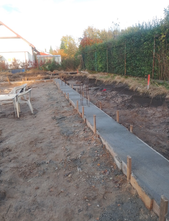 Świeżo wylany, długi fundament pod budowę domu, z wystającymi prętami zbrojeniowymi i drewnianym szalunkiem, na tle ogrodu i domu jednorodzinnego w pochmurny dzień.