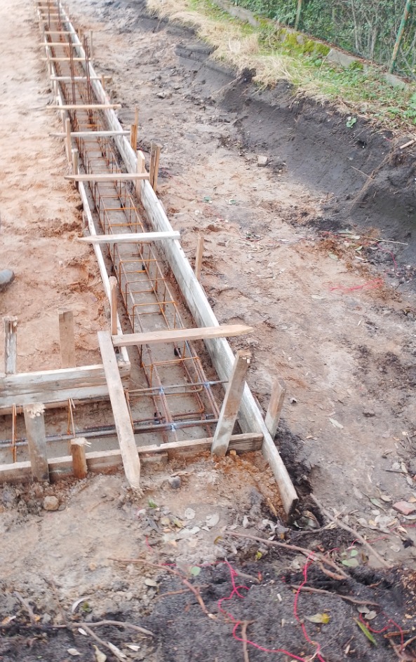 Przygotowanie szalunku i zbrojenia pod fundament liniowy, widoczne drewniane deski i stalowe pręty zbrojeniowe, ziemia i trawa w tle.