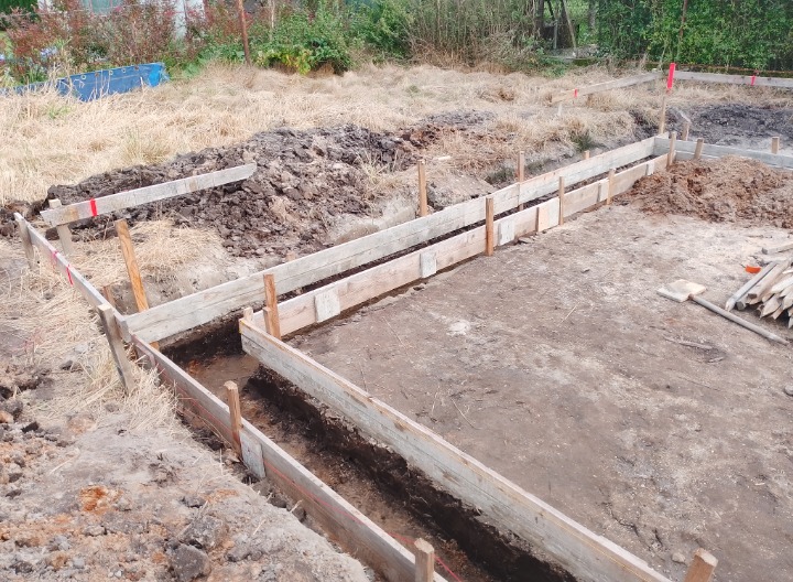 Wykonanie szalunku pod fundamenty z drewnianych desek, widoczne wykopane rowy, pionowe pale podtrzymujące konstrukcję i ziemia usypana obok wykopu.