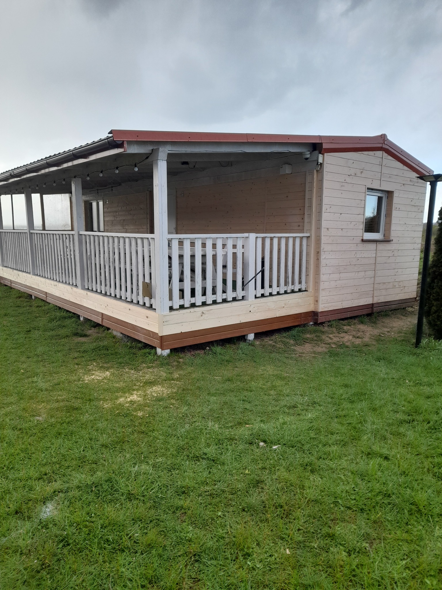 Drewniany domek letniskowy z zadaszonym tarasem, pomalowany na jasny kolor z brązowymi akcentami, otoczony trawnikiem, w pochmurny dzień.