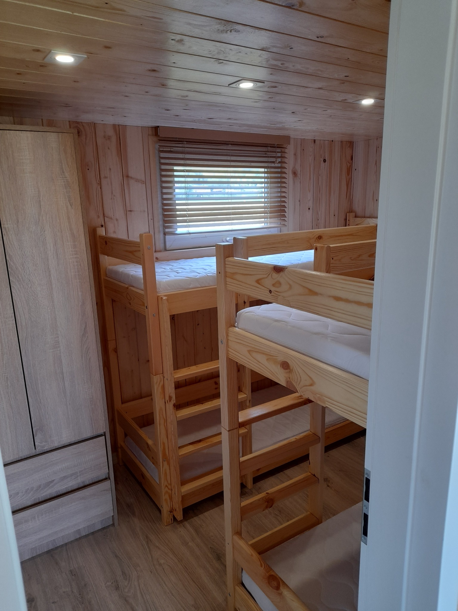 Drewniane wnętrze z łóżkiem piętrowym, szafą i oknem z żaluzjami, oświetlone punktowymi lampami sufitowymi.