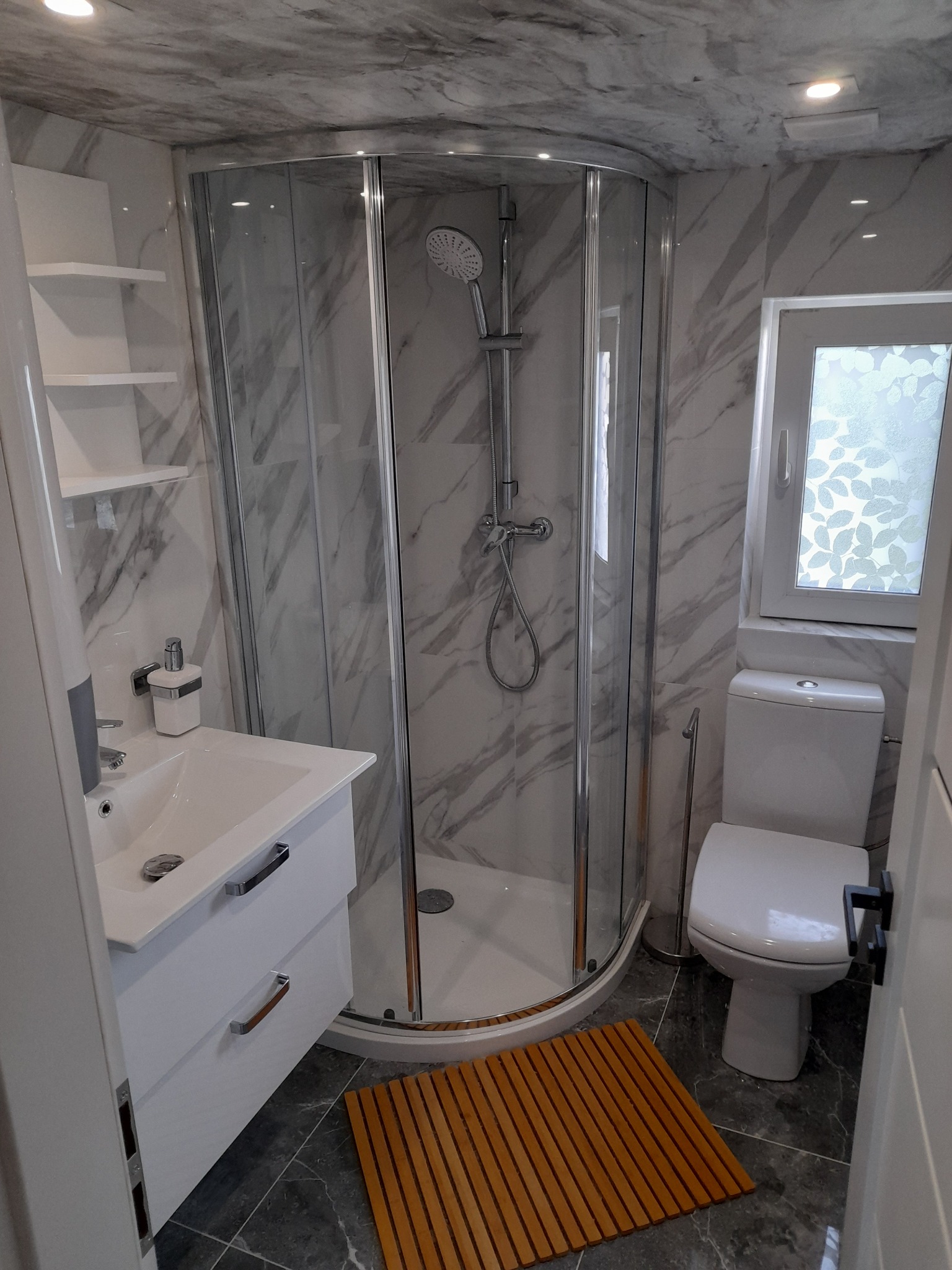 Nowoczesna łazienka z marmurowymi płytkami, kabiną prysznicową z zaokrąglonymi drzwiami, białą umywalką z szafką, toaletą i drewnianą wycieraczką na podłodze.