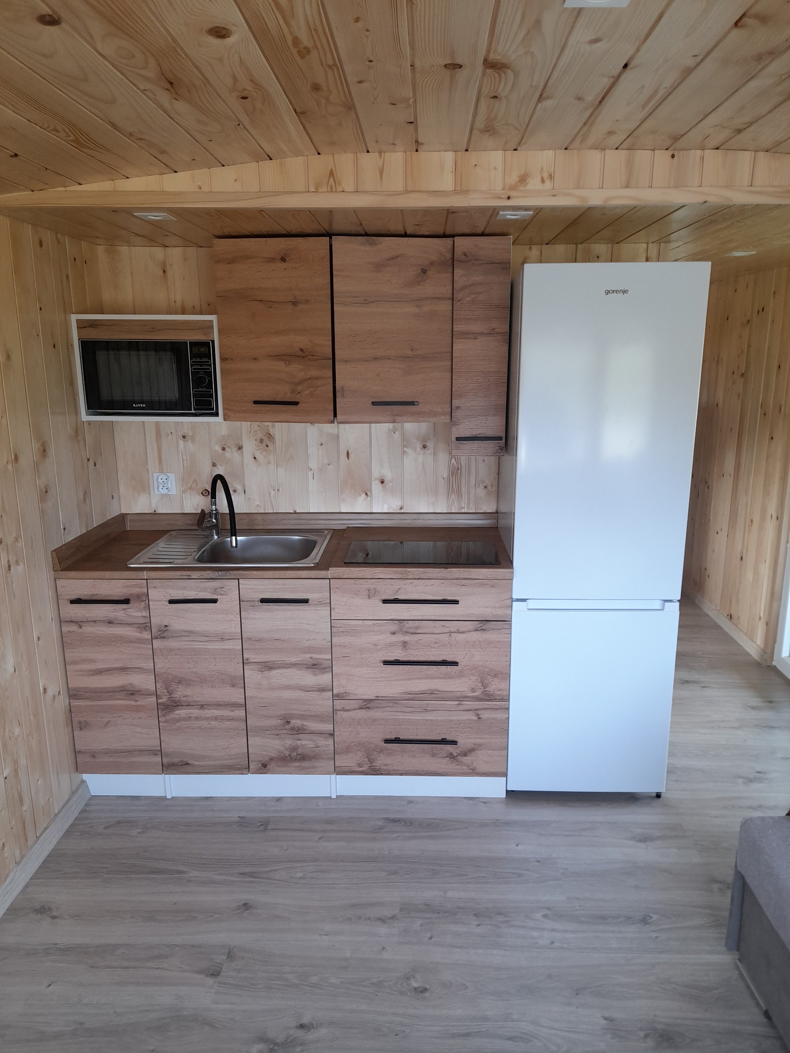 Drewniana kuchnia w domku letniskowym: zabudowa z drewnianymi frontami, biała lodówka Gorenje, mikrofalówka Raven, zlewozmywak ze stali nierdzewnej i czarna bateria.