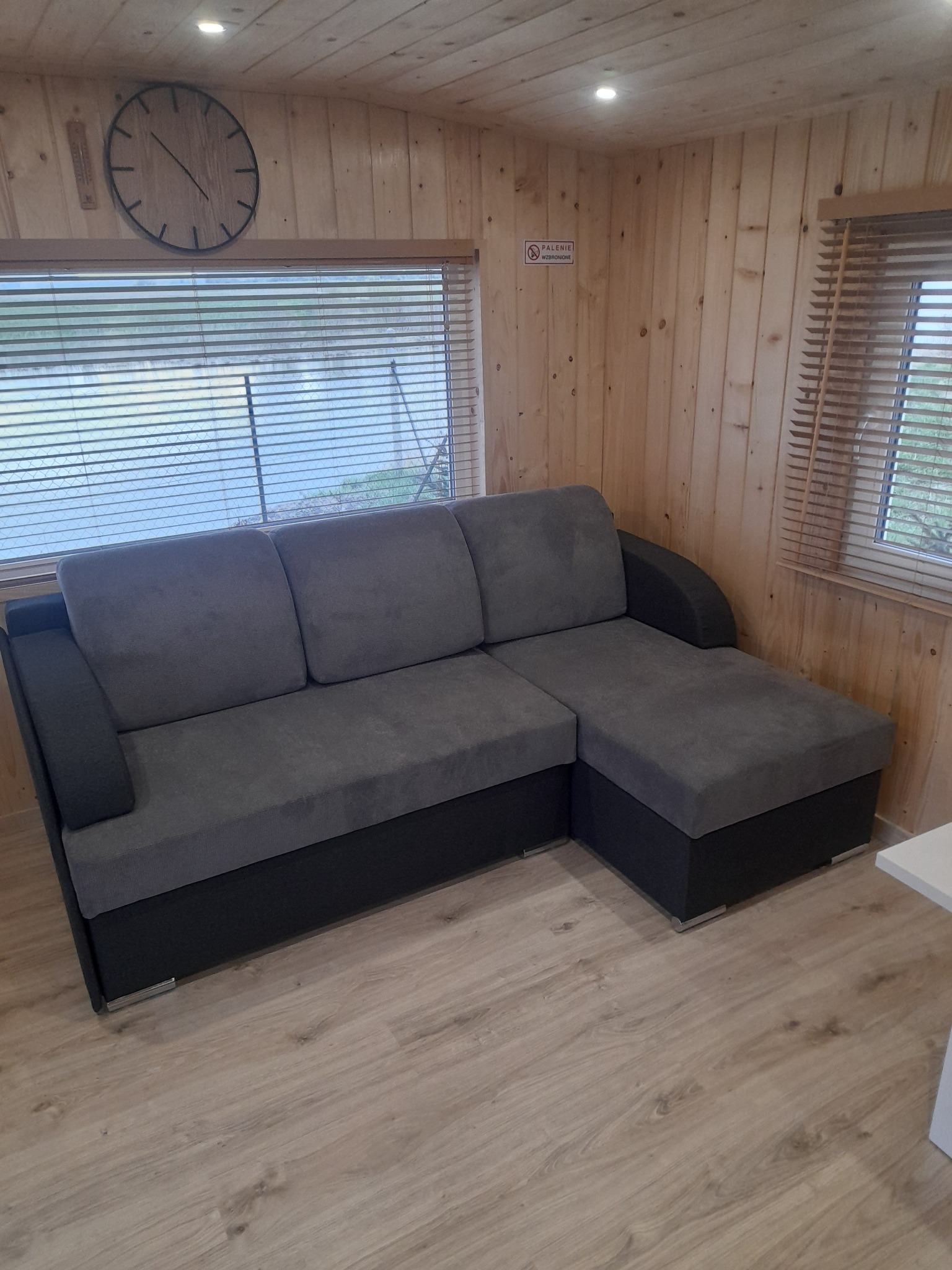 Szara sofa narożna z czarną podstawą w pomieszczeniu wykończonym drewnianą boazerią, widok na zewnątrz przez okno z żaluzjami, zegar na ścianie.