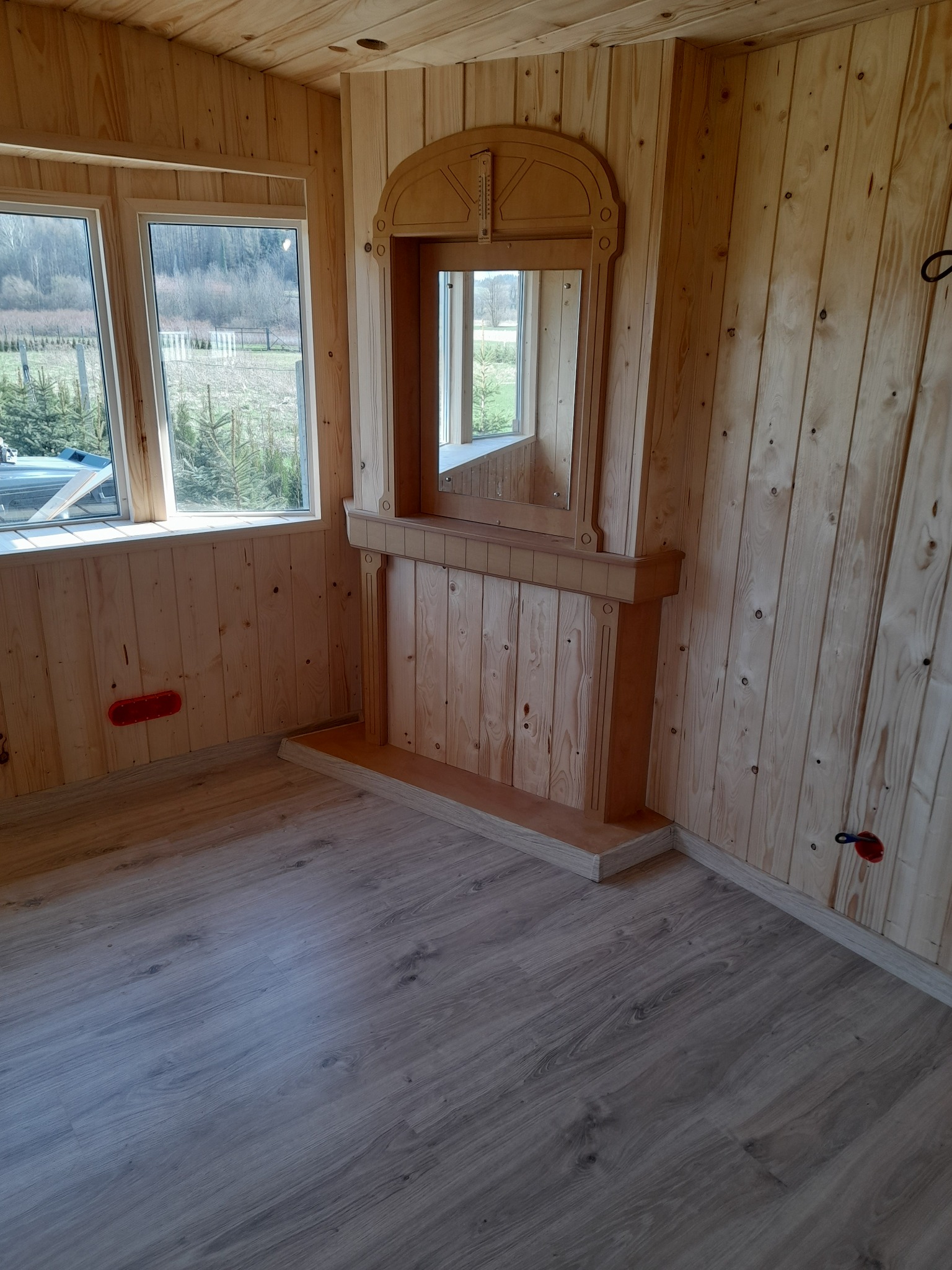 Wnętrze pomieszczenia wykończonego boazerią z jasnego drewna, z drewnianą atrapą kominka z lustrem i widokiem na zewnątrz przez okno.