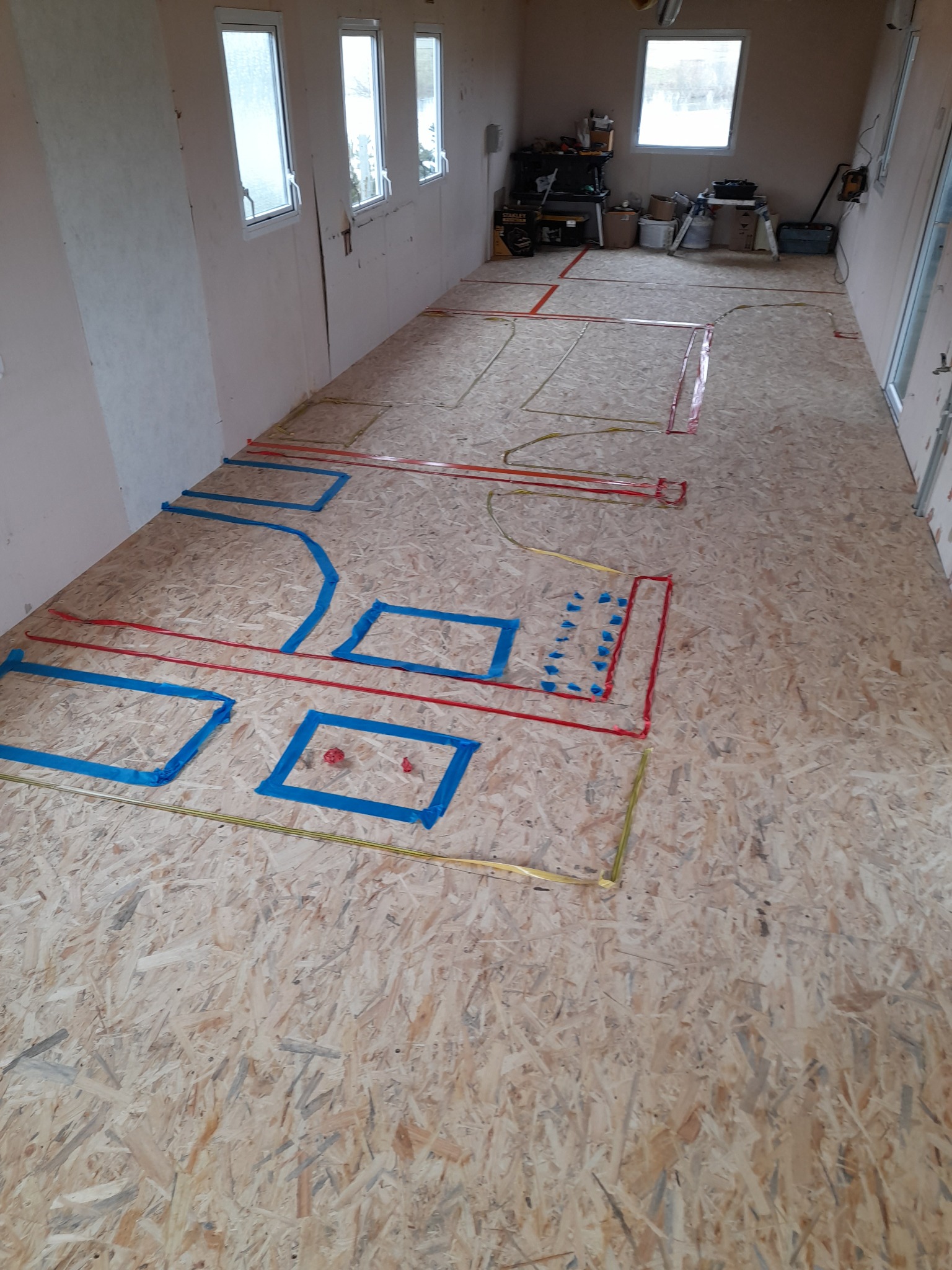 Przygotowanie podłoża z płyty OSB pod instalacje elektryczne i ogrzewanie podłogowe, oznaczone kolorowymi taśmami w długim pomieszczeniu z oknami i narzędziami w tle.
