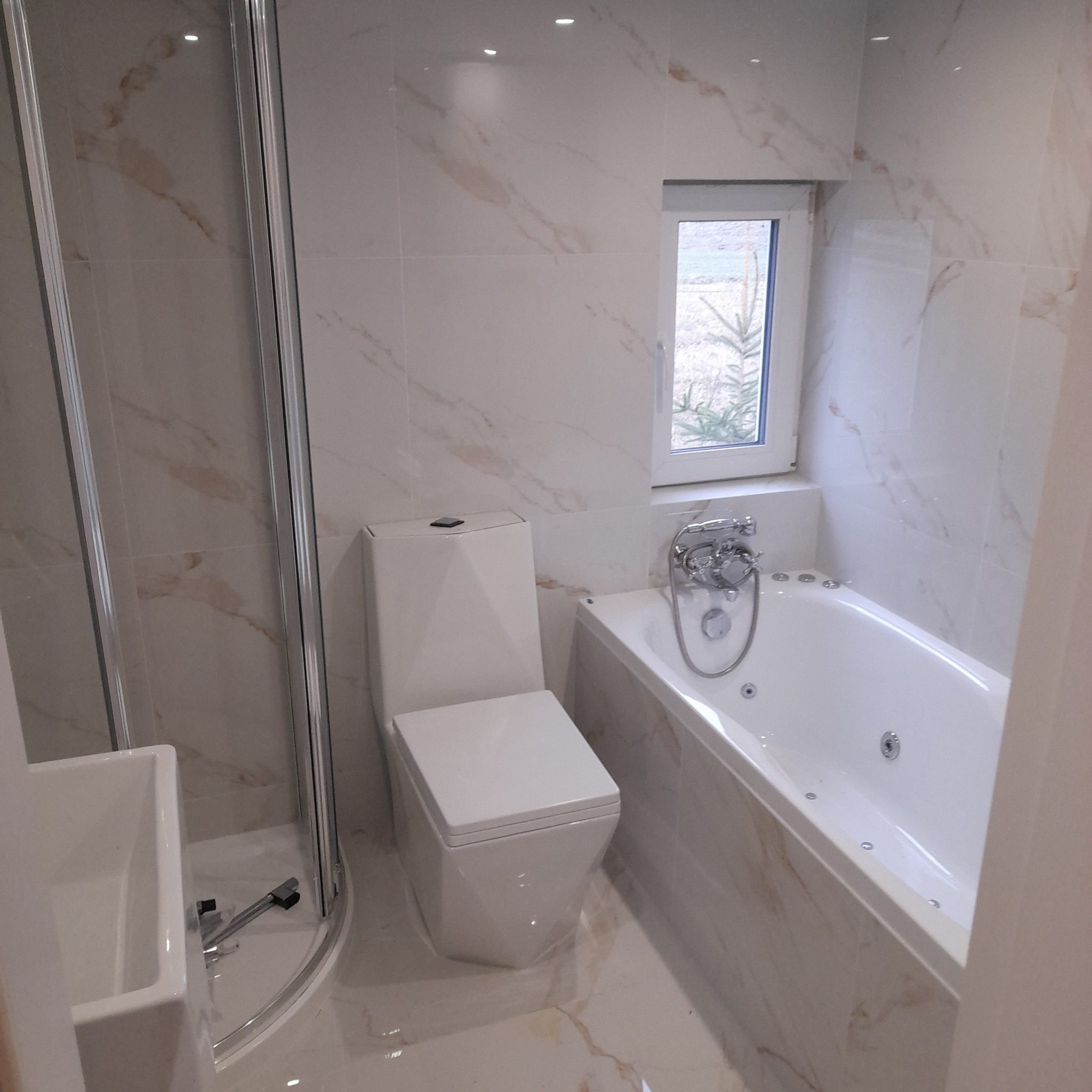 Nowoczesna łazienka z beżowymi płytkami imitującymi marmur, kabiną prysznicową z chromowanymi profilami, białą toaletą i wanną z hydromasażem pod oknem.