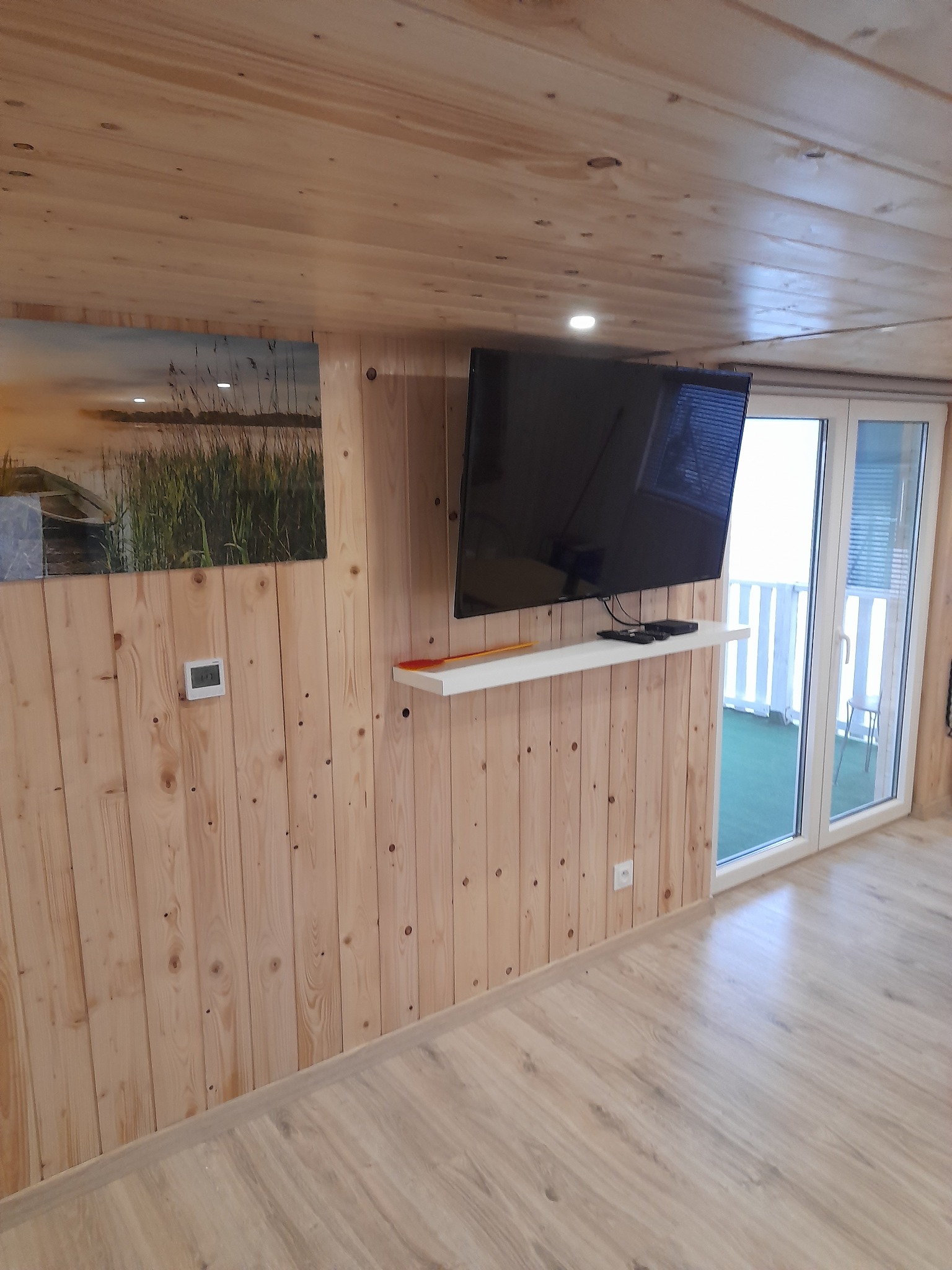 Wnętrze balkonu z drewnianą boazerią, telewizorem na ścianie, obrazkiem z motywem jeziora i wyjściem na balkon z białymi barierkami.