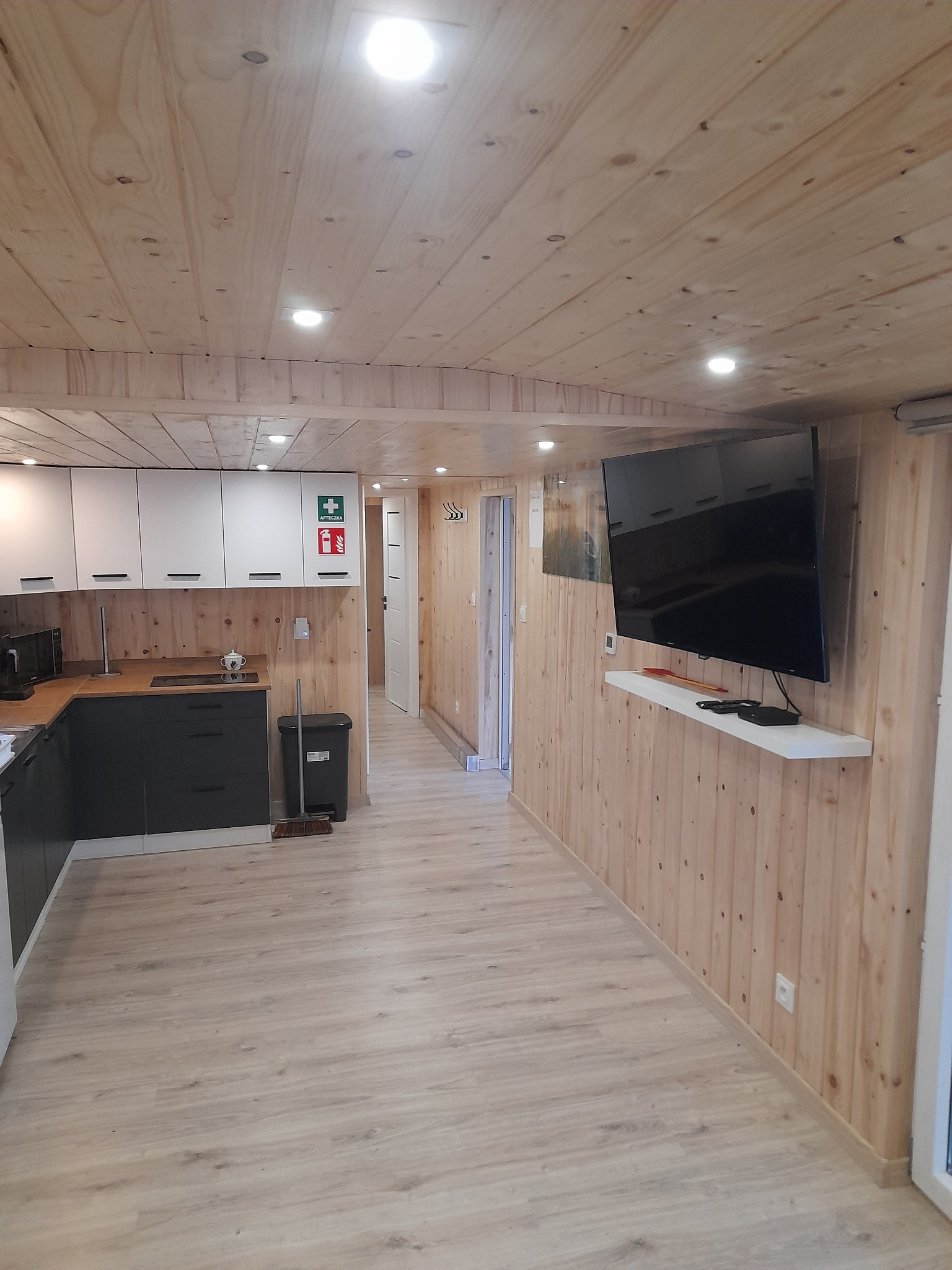 Wnętrze wykończone drewnem: ściany, sufit, podłoga laminowana, czarno-biała zabudowa kuchenna, telewizor na ścianie, apteczka na szafce.
