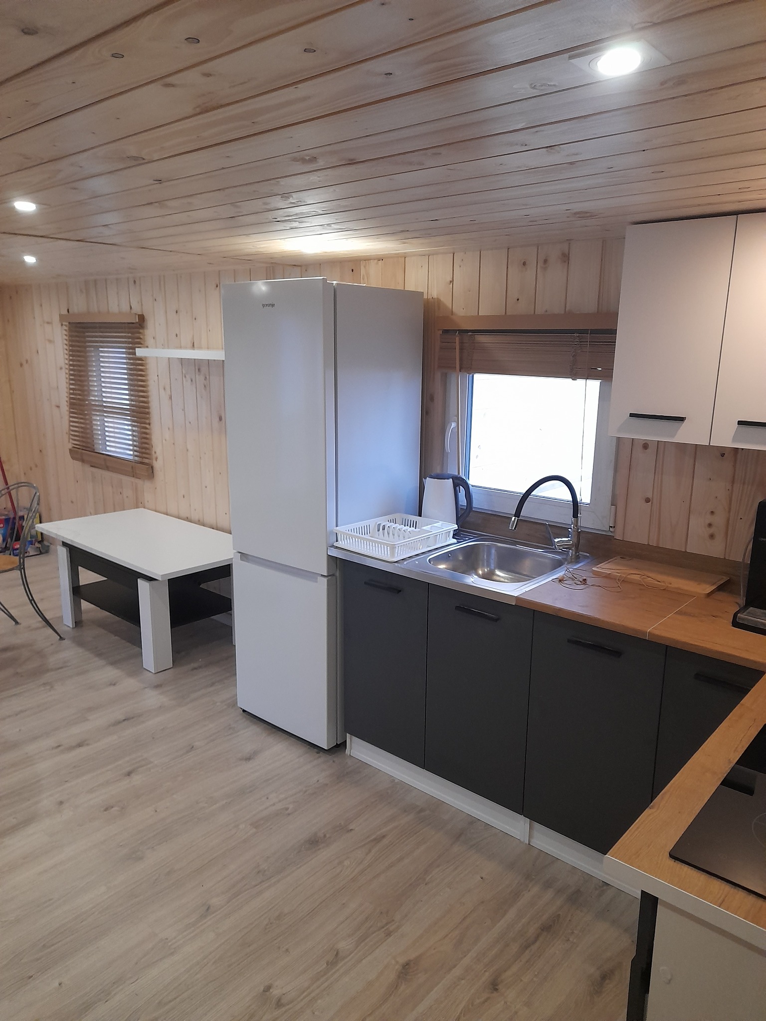 Wnętrze kuchni z drewnianymi ścianami, zabudową kuchenną w odcieniach szarości i bieli, lodówką Gorenje oraz stołem z krzesłem w tle.