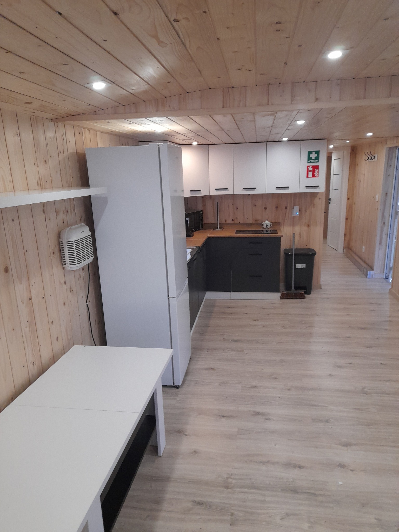 Wnętrze małego domku z drewnianymi ścianami i sufitem, wyposażonego w białą lodówkę, biało-czarne szafki kuchenne, biały stół i grzejnik na ścianie.