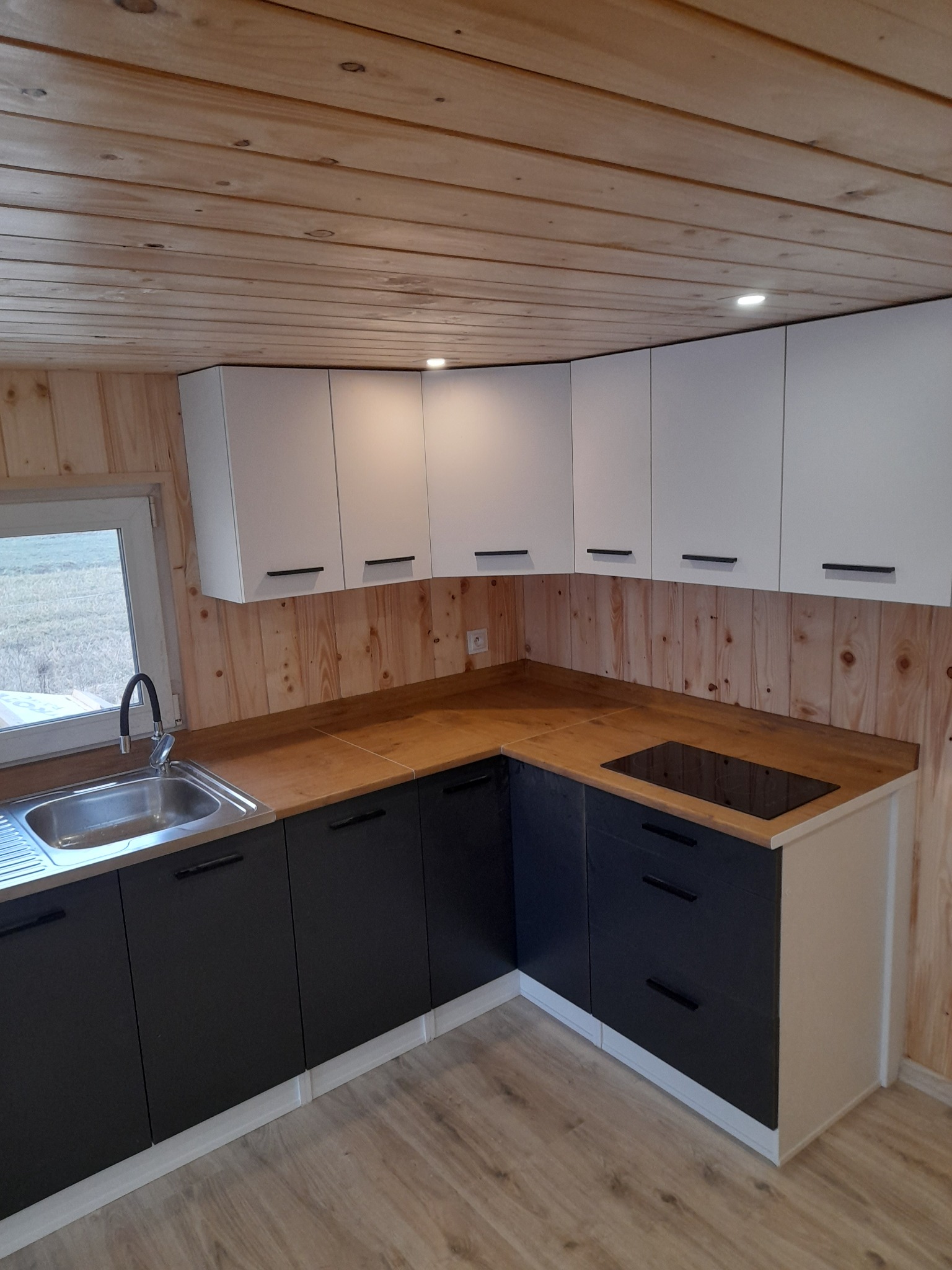 Nowoczesna kuchnia w domku z drewnianym wykończeniem, białe szafki górne z czarnymi uchwytami, ciemne szafki dolne, drewniany blat i płyta indukcyjna.