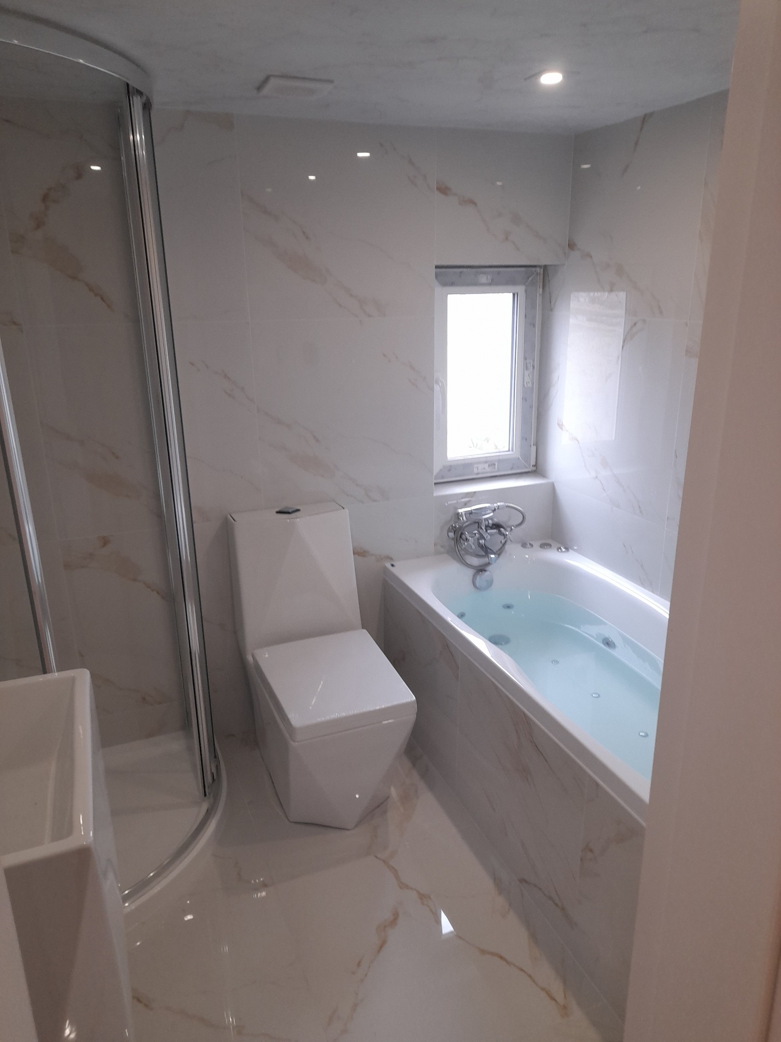 Nowoczesna łazienka z marmurowymi płytkami, białą wanną z hydromasażem, toaletą typu compact i kabiną prysznicową z chromowanymi profilami.