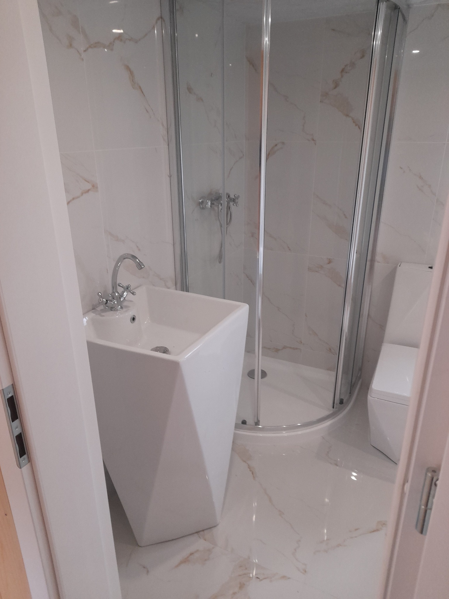 Biała, geometryczna umywalka wolnostojąca z chromowaną baterią w narożnej kabinie prysznicowej wyłożonej marmurowymi płytkami z brązowymi żyłami, widok z poziomu podłogi.