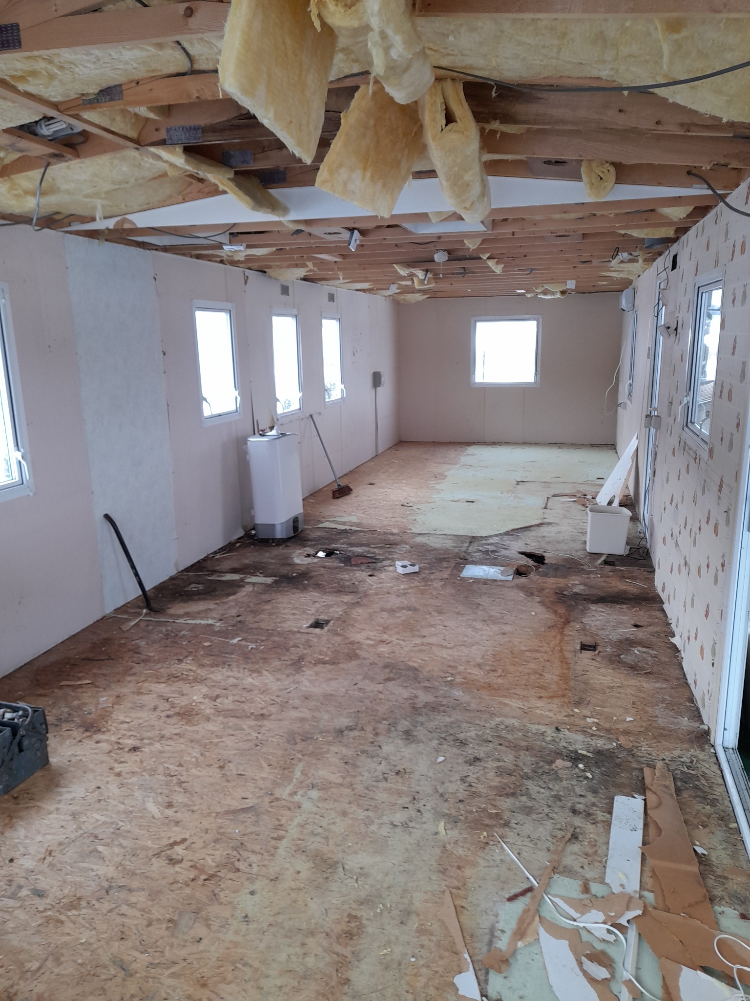 Wnętrze w trakcie demontażu: odsłonięta konstrukcja sufitu z izolacją, goła podłoga z płyty OSB, ściany częściowo z tapetą, widoczne okna, grzejnik i narzędzia.