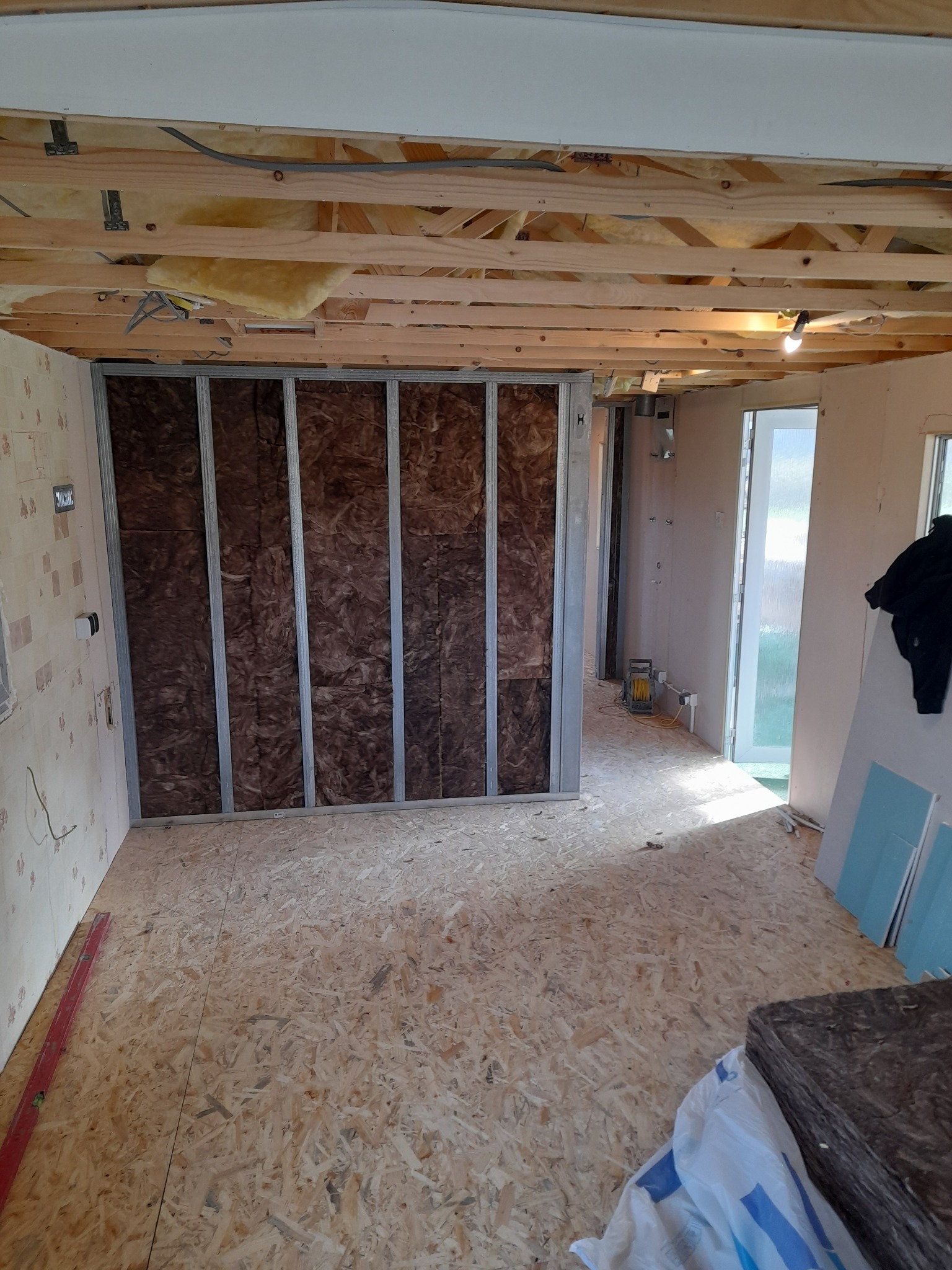 Wnętrze w trakcie adaptacji: widoczna konstrukcja metalowa ściany działowej z izolacją z wełny mineralnej, surowa podłoga z płyt OSB, odsłonięty strop z widocznymi elementami instalacji elektrycznej.