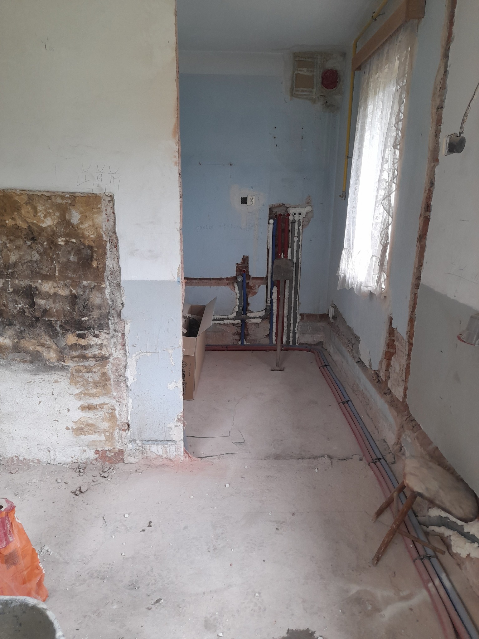 Wnętrze łazienki w trakcie remontu: odsłonięte instalacje wodne, fragmenty starej glazury, resztki tynku na ścianach, prowizoryczne siedzisko i kartonowe pudło w przejściu do kolejnego pomieszczenia.