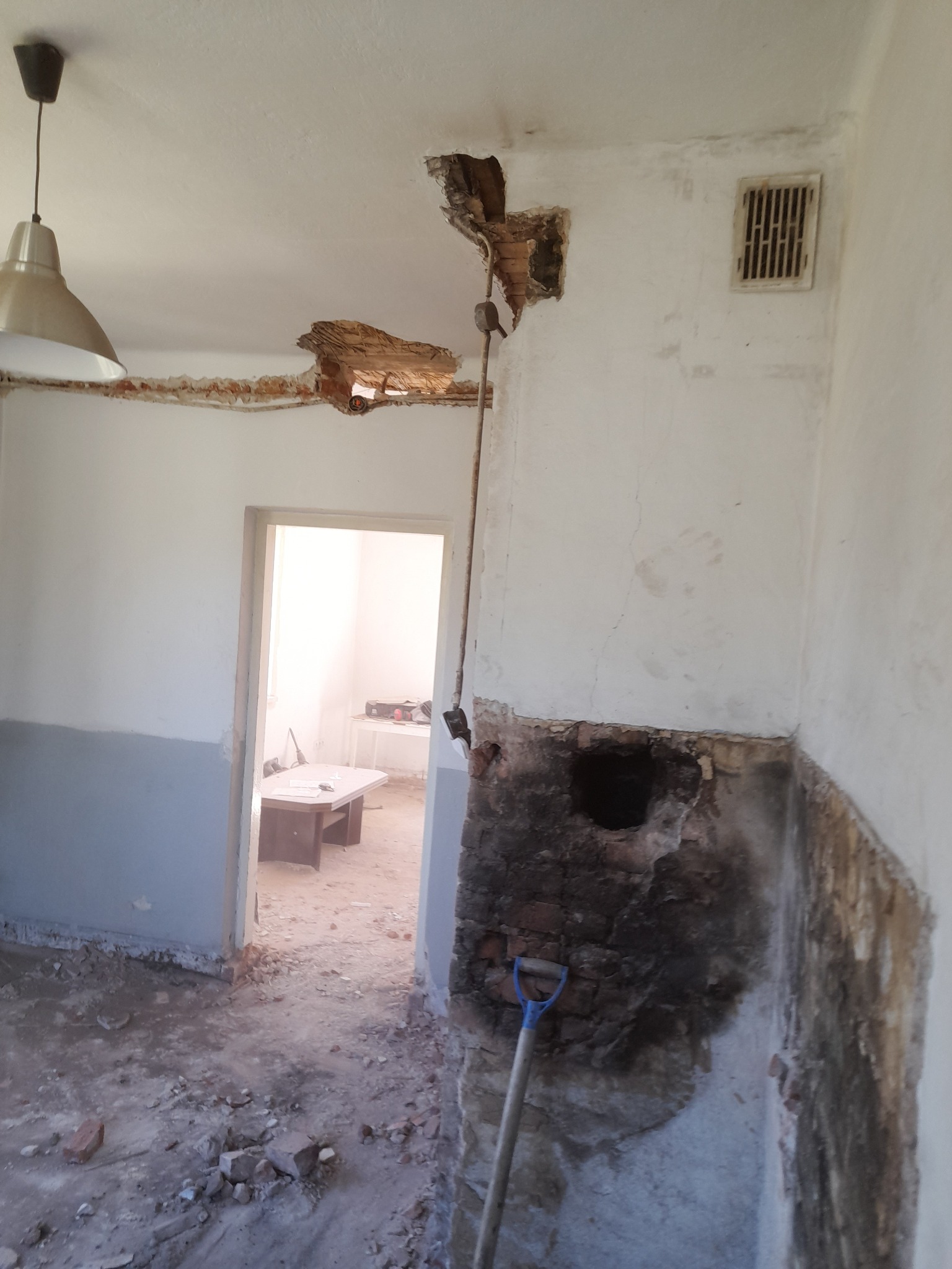 Pomieszczenie w trakcie prac przygotowawczych do remontu, widoczne uszkodzenia ścian i sufitu, gruz na podłodze, oraz łopata oparta o ścianę, sugerujące prace rozbiórkowe.