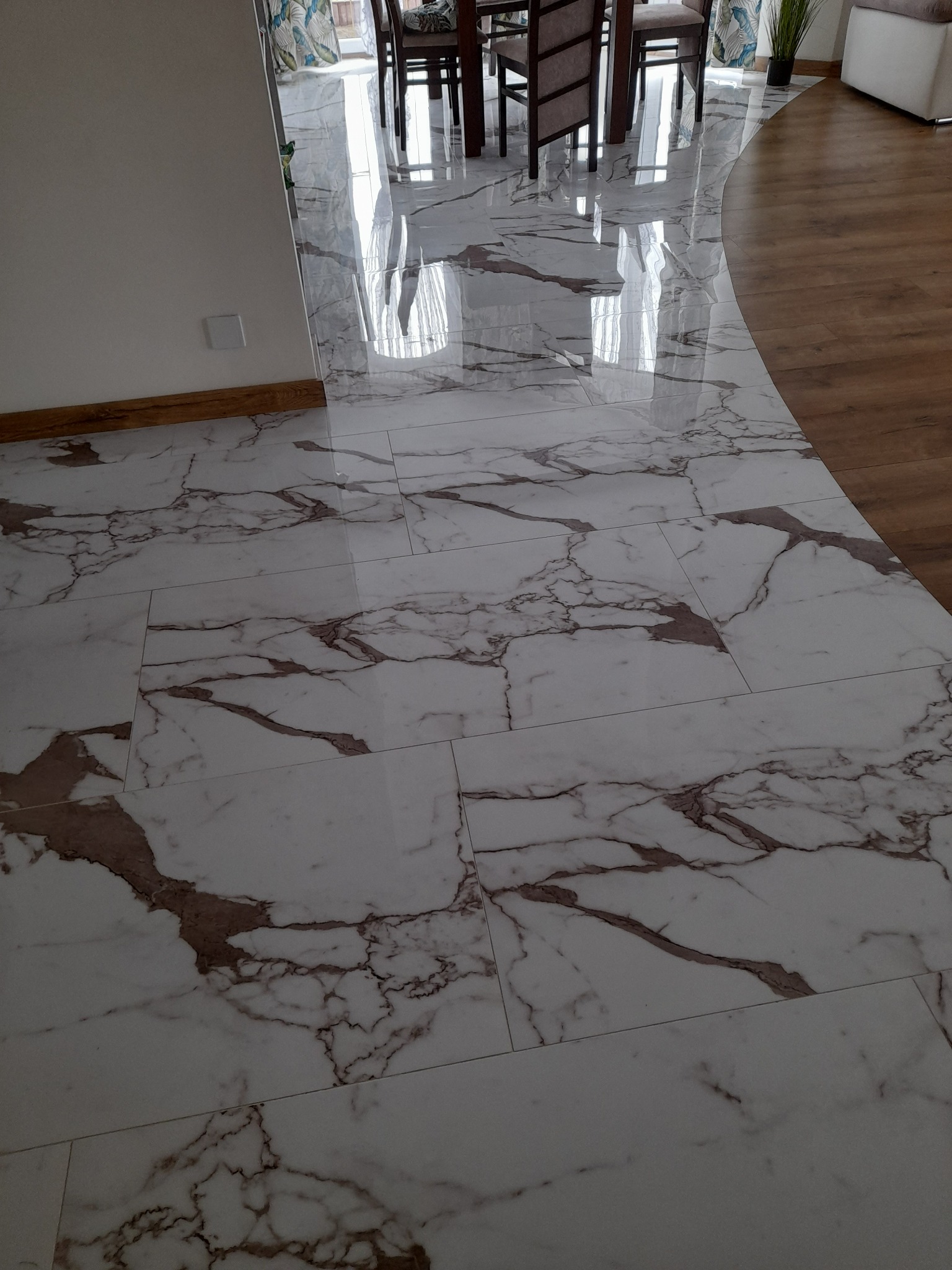 Błyszcząca podłoga z płytek imitujących marmur Carrara, z widocznym przejściem na drewniany parkiet w tle, odbijająca światło z okna.