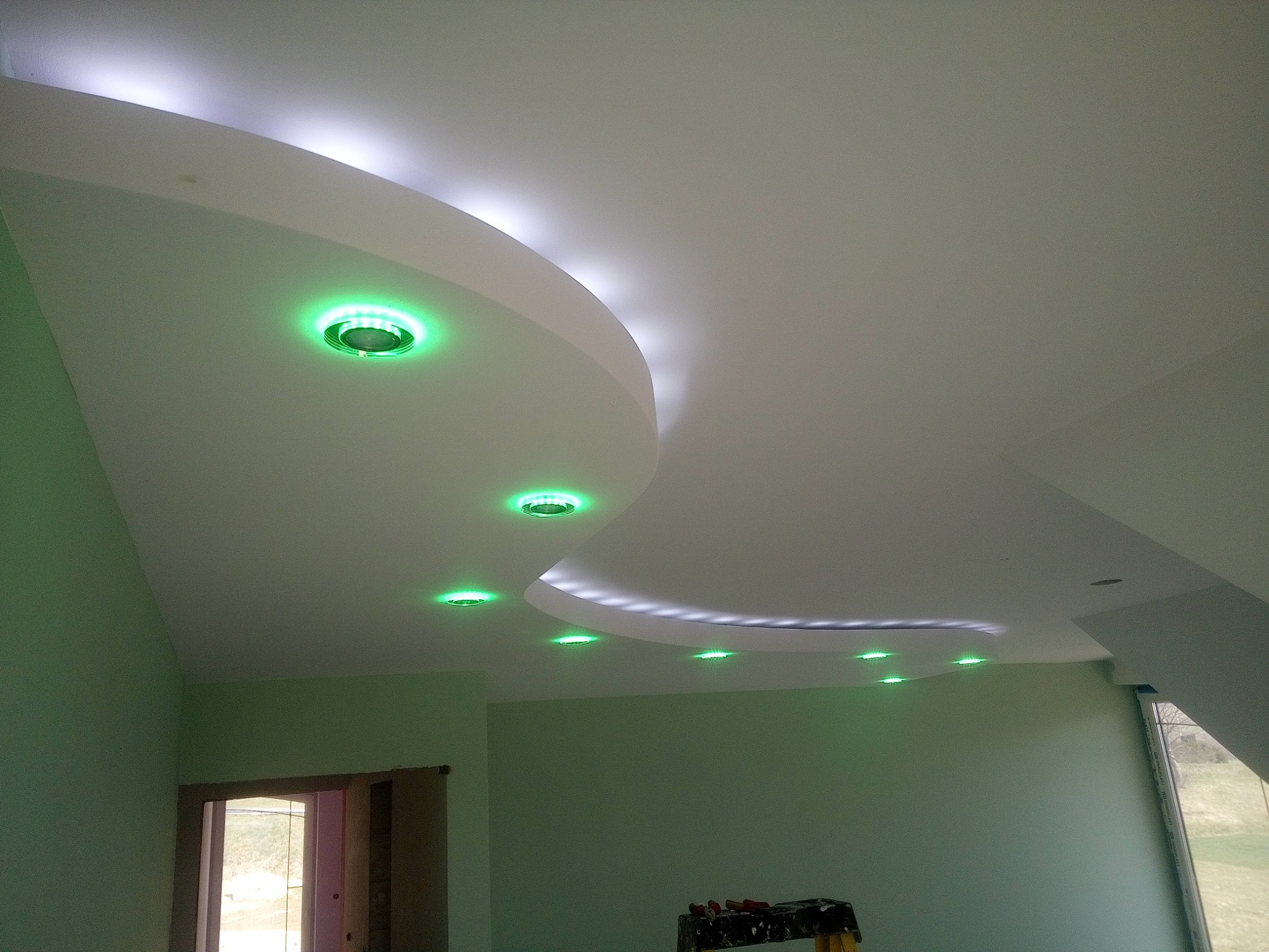 Podwieszany sufit z dekoracyjnym oświetleniem LED w kolorze zielonym i białym, świeżo pomalowane ściany w odcieniach zieleni, drabina malarska w rogu pokoju.